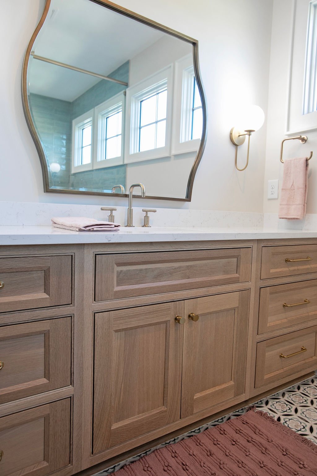 Bathroom vanity with a large mirror, light fixture, two towels, wooden cabinetry, pink rug, and patterned floor tiles.