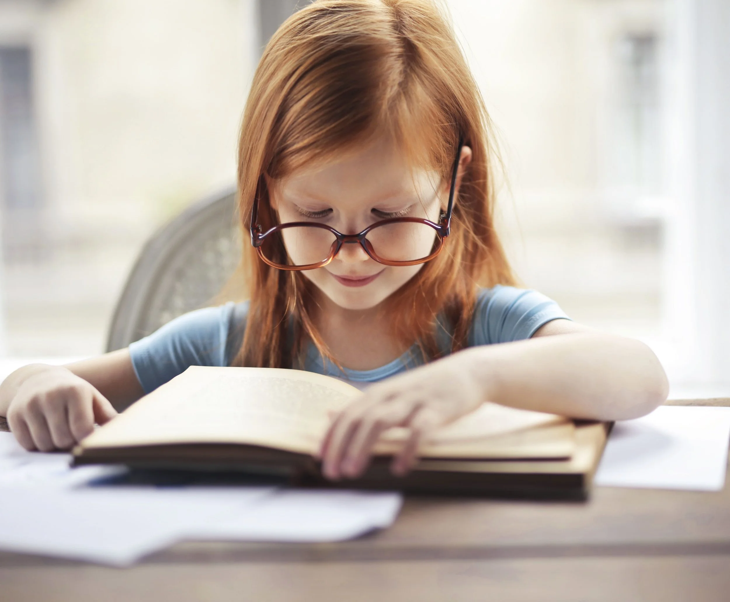 A young girl wearing glasses is concentrating on a book.