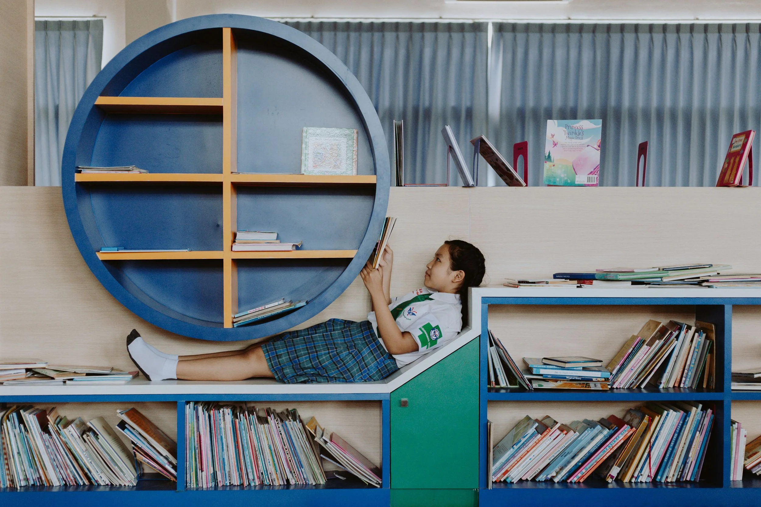 A young girl in a school uniform reclines against a bookshelf and reads in a library.
