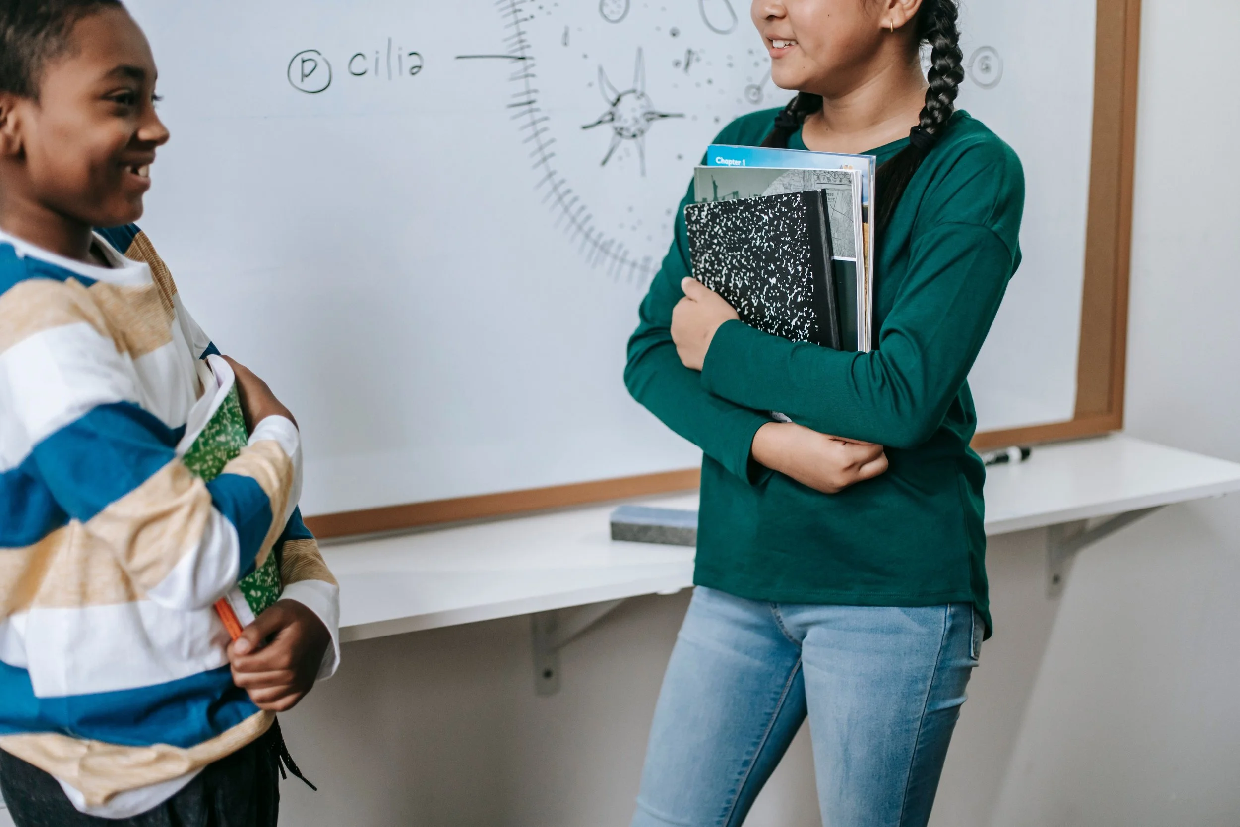two happy children are facing each other in front of a whiteboard and holding excercise books. On the whiteboard is a diagram of a cell. Both children look relaxed and confident.