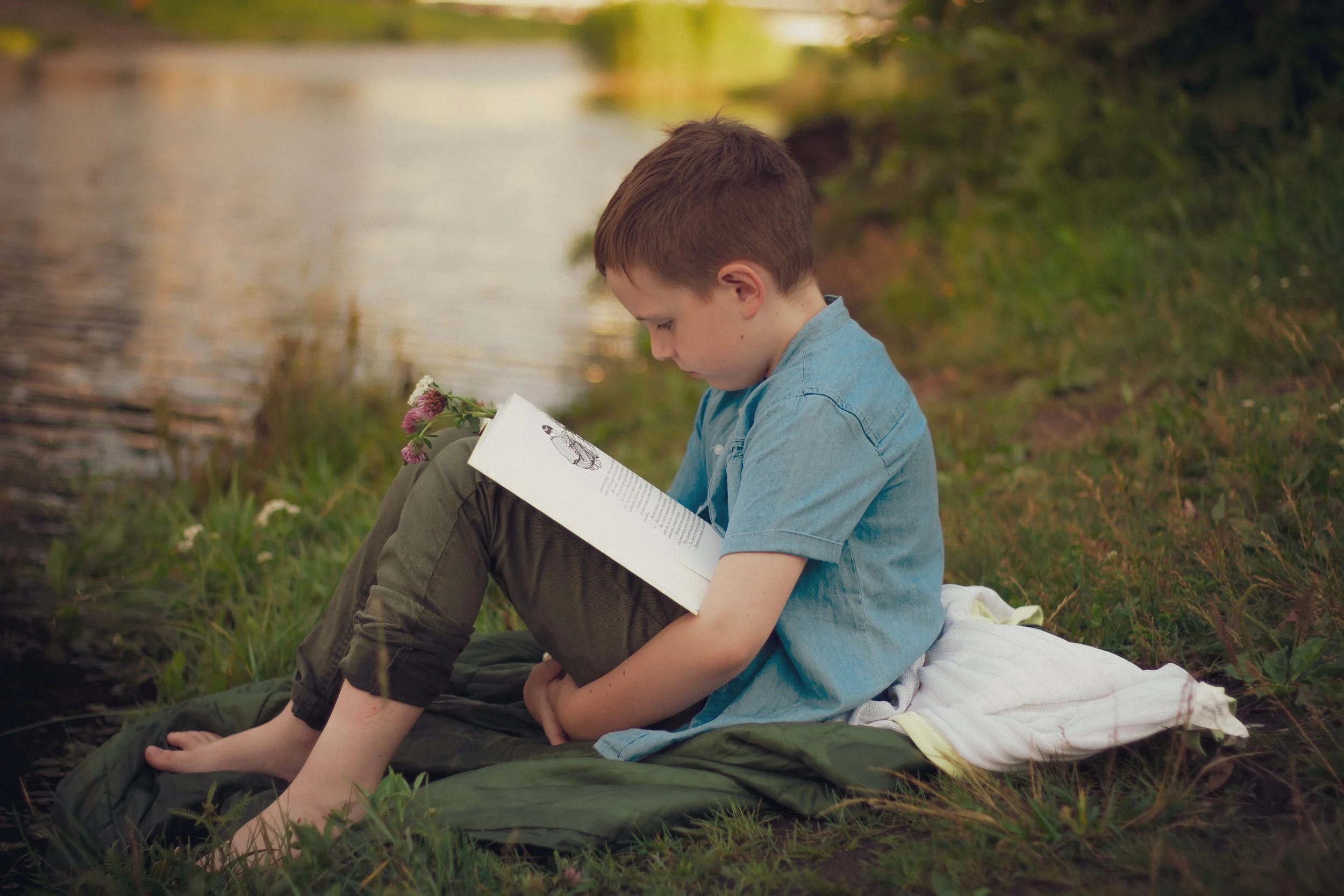 A young boy reading, concentrating on the book, by the riverbank on a sunny day.