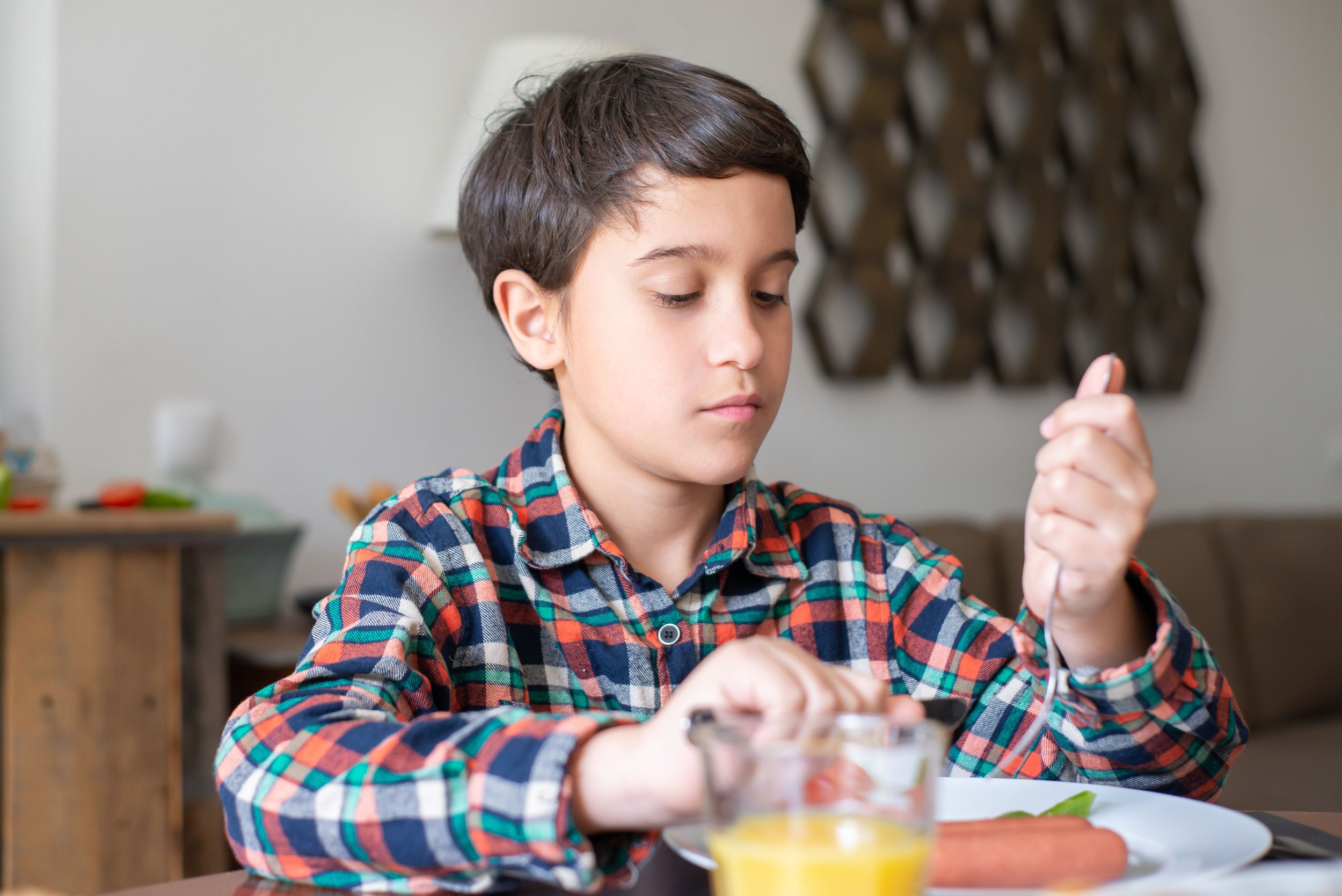 A boy sits at a kitchen table holding cutlery looks thoughtfully at his plate, unsure what to eat first.