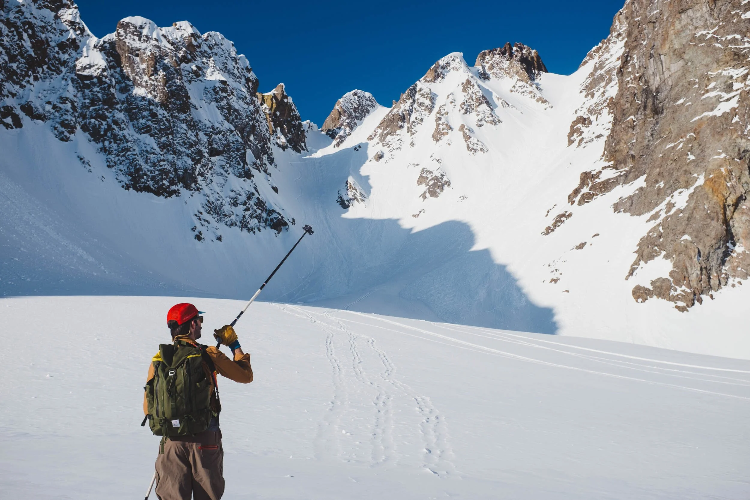 Two skiers in bright clothing standing on a snowy mountain slope, with rugged snow-covered peaks and a clear blue sky in the background.