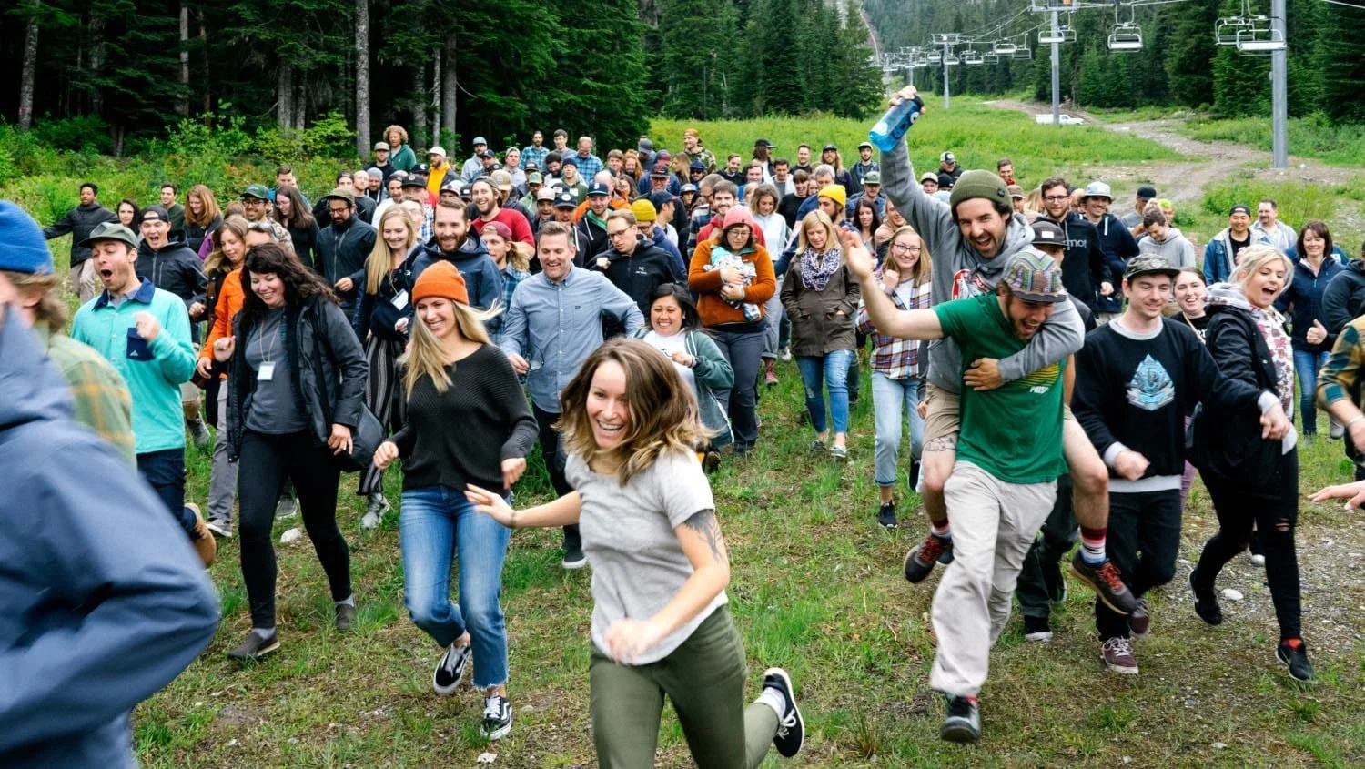 Large group of cheerful people running outdoors on grassy terrain in a forested area, some are smiling and laughing, with a ski lift visible in the background.