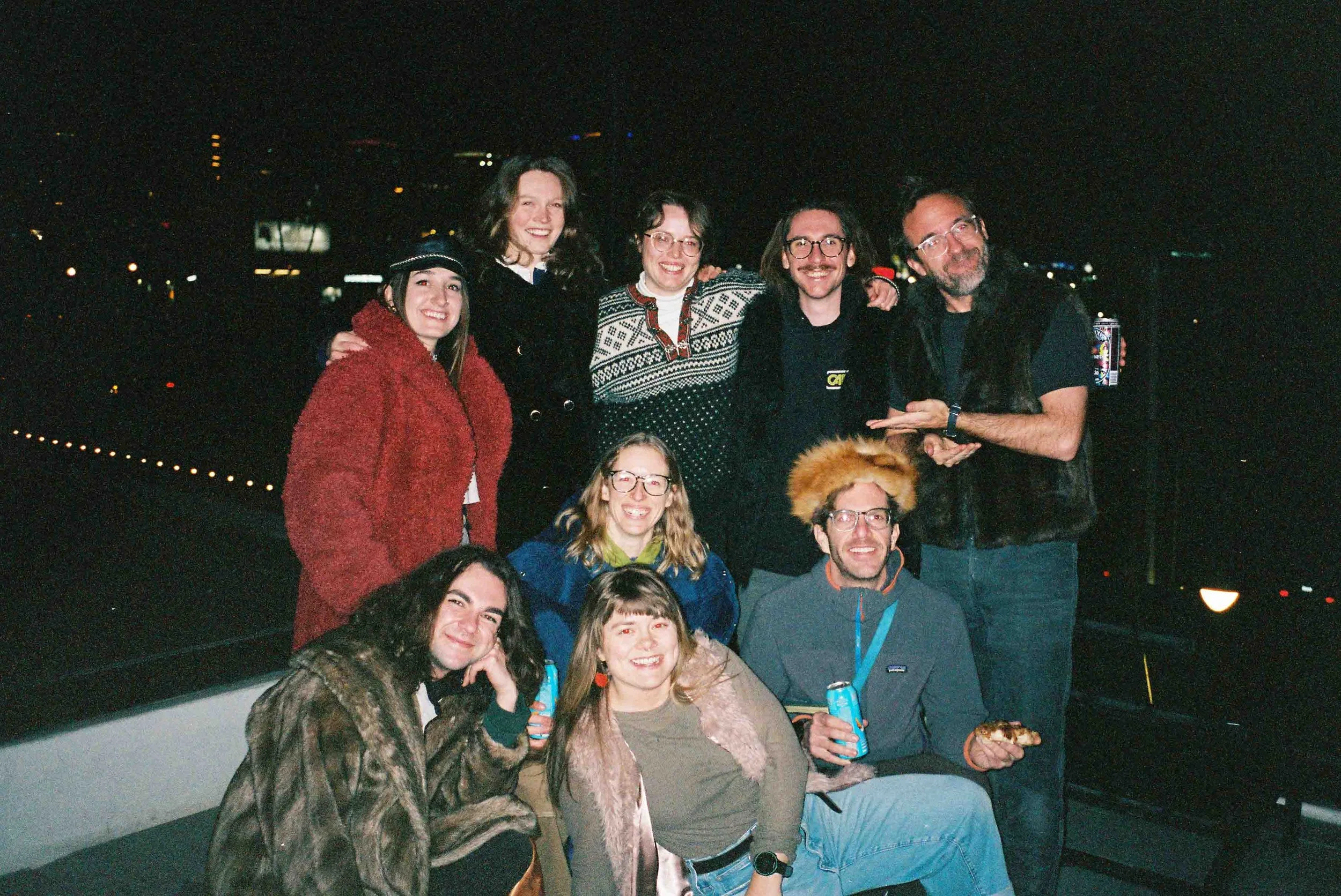 Group of people, mostly young adults, smiling at night on a rooftop with city lights in the background, holding drinks and pizza.