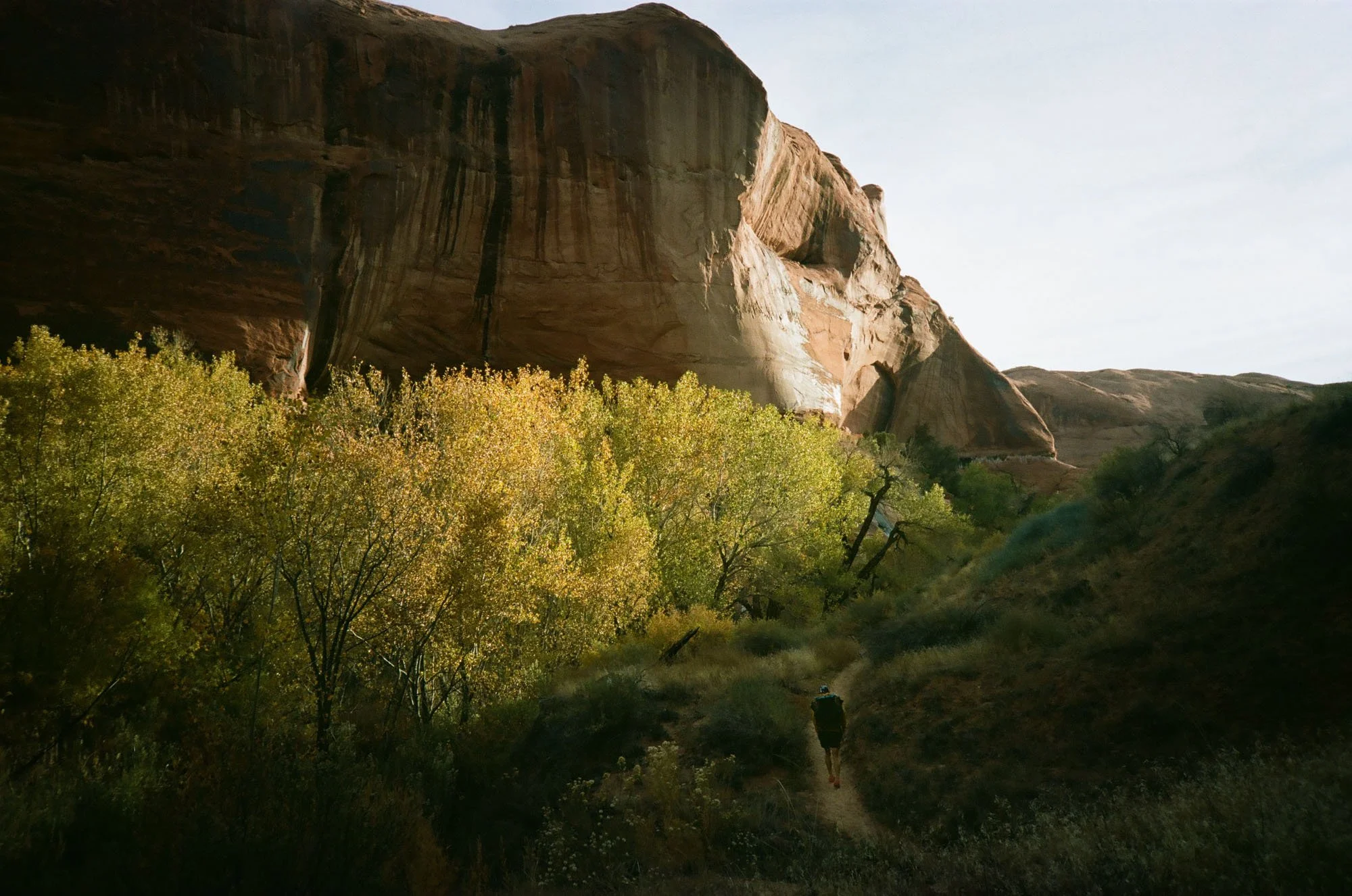 A person walking on a trail in a mountainous landscape with a large rock formation and trees with yellow foliage, under a clear sky.