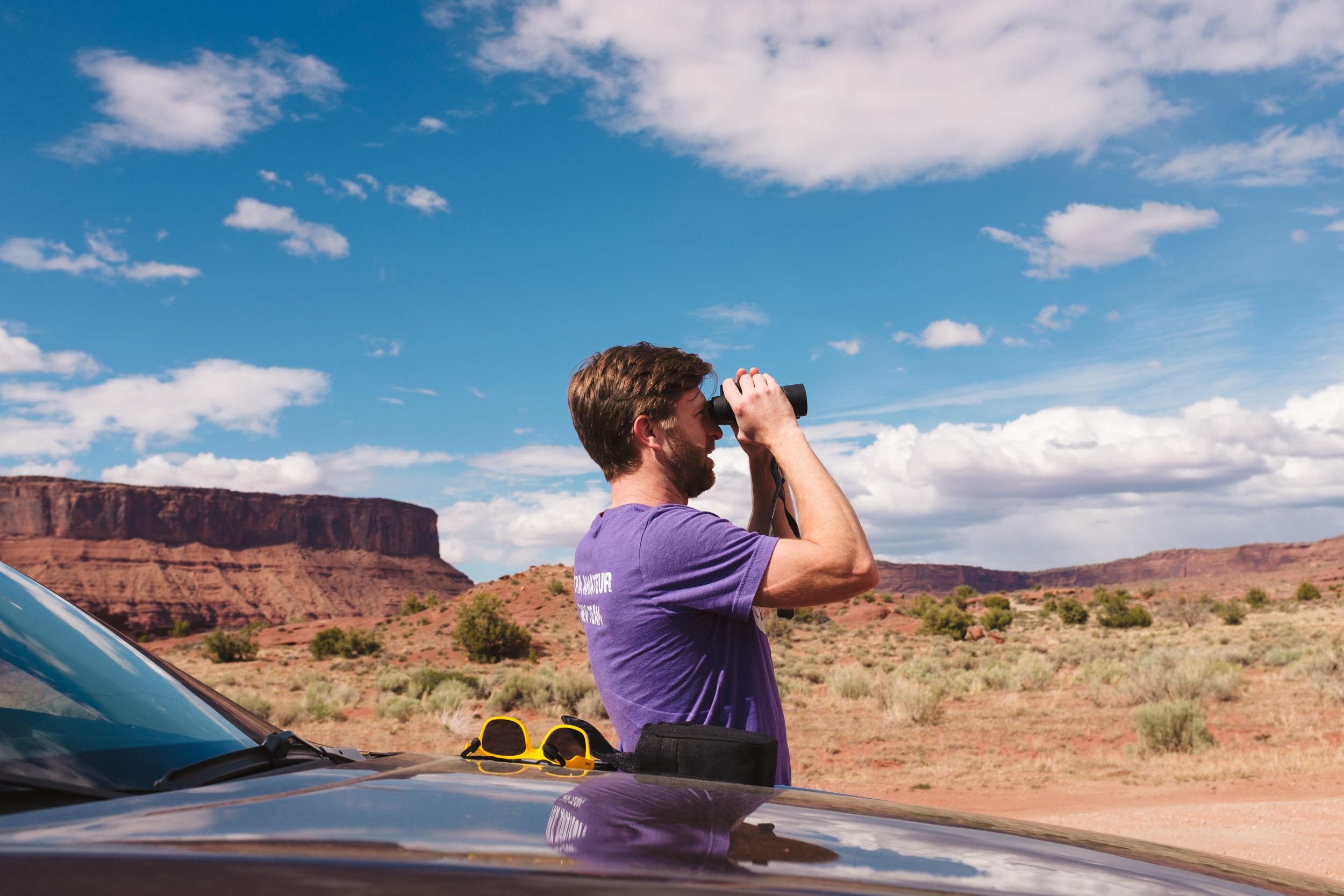A man with brown hair and a beard, wearing a purple t-shirt, is standing outdoors looking through binoculars at a desert landscape with red rock formations and a blue sky with white clouds.