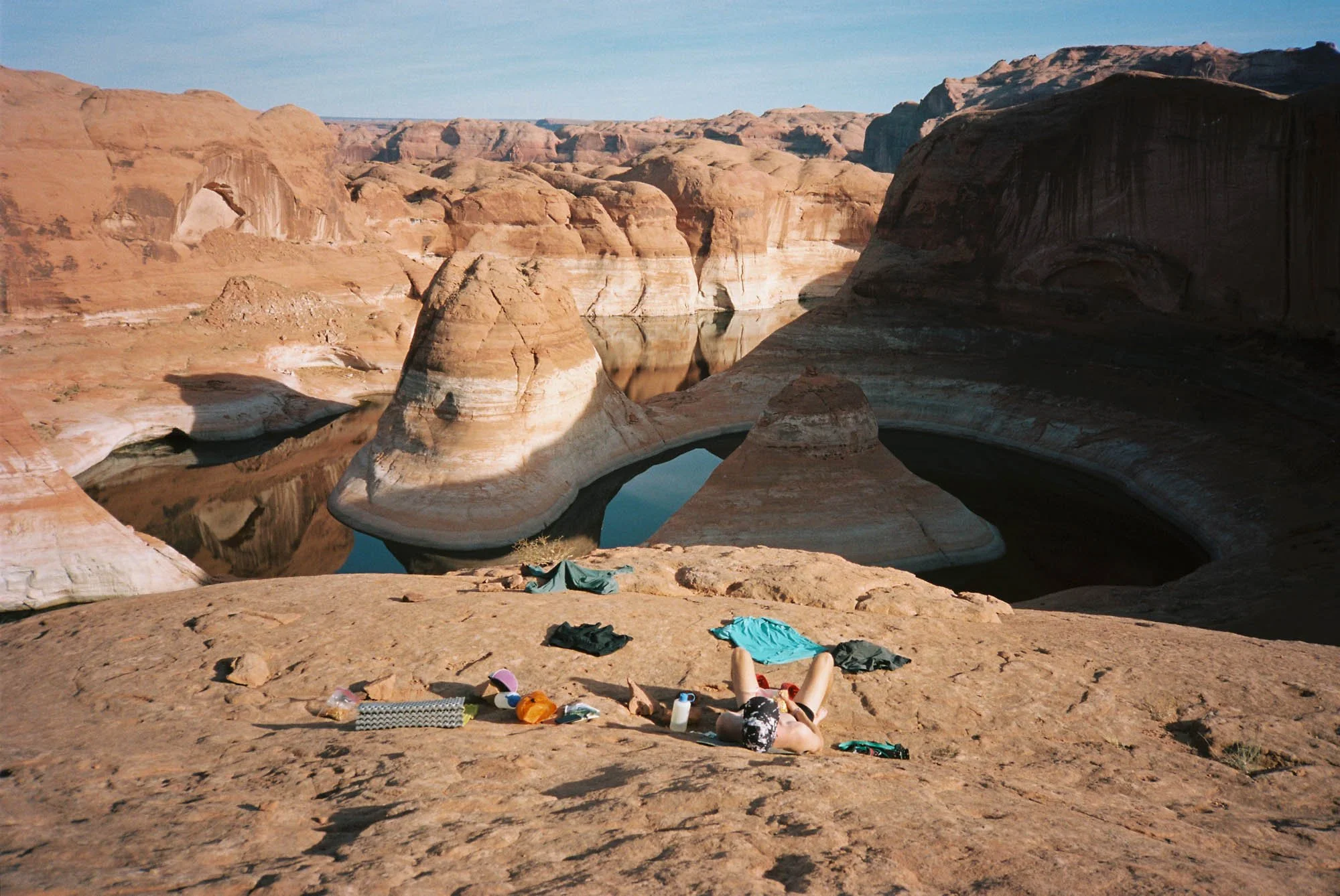 A desert landscape with red rock formations and a body of water reflecting the cliffs. The foreground has scattered clothing and personal items, with a person lying on the ground.