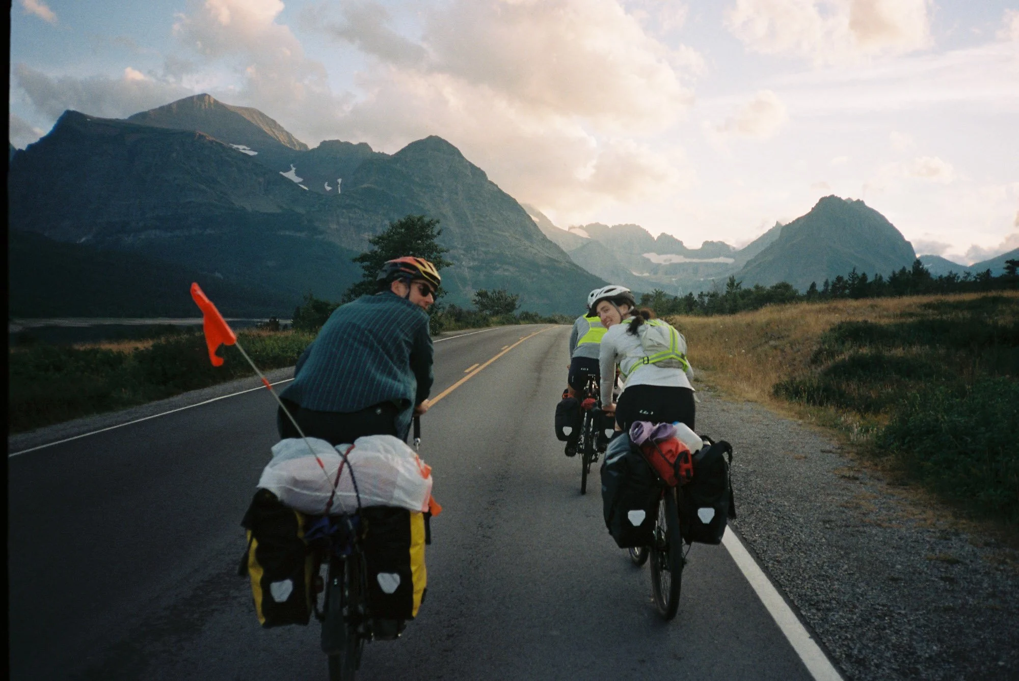 Group of people biking along a scenic mountain road, with mountains and cloudy sky in the background.