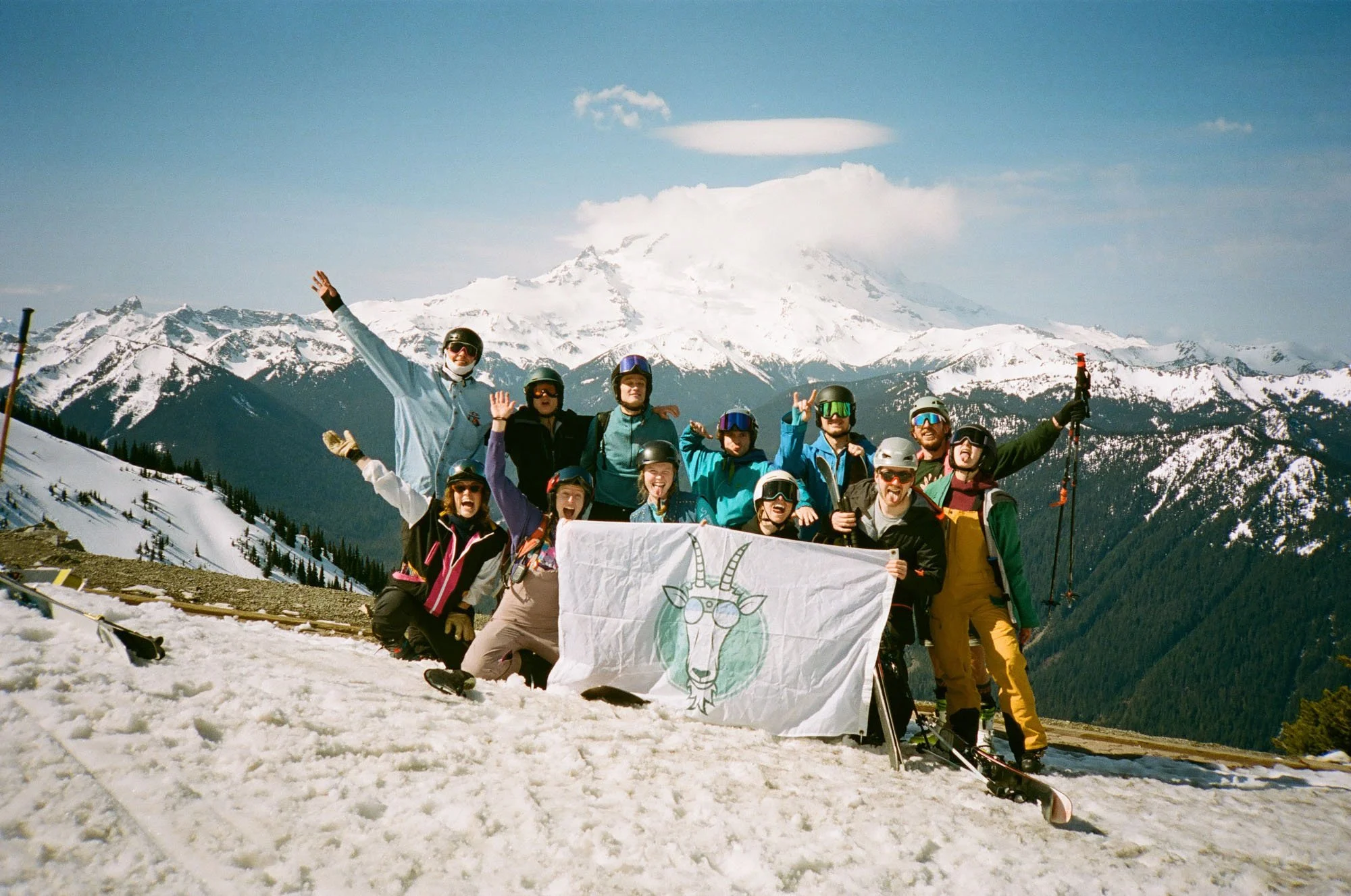 Group of skiers and snowboarders on a snowy mountain with snow-capped peaks and a volcano in the background, holding a white flag with a cartoon llama wearing glasses.