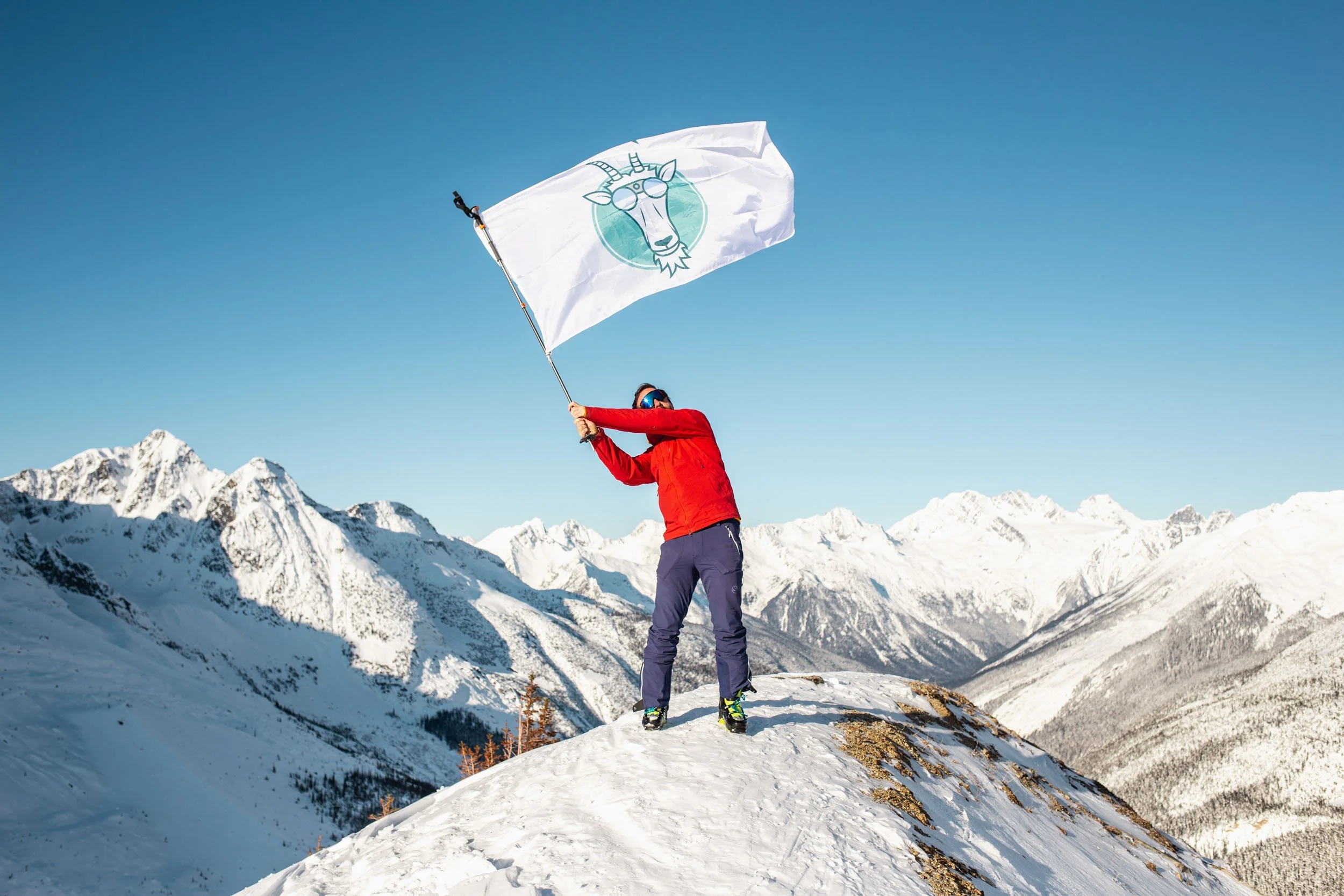 A person dressed in a red jacket, blue pants, and snow boots holding a flag with a goat head logo, standing on snowy mountain terrain with a backdrop of snow-covered mountains and a clear blue sky.
