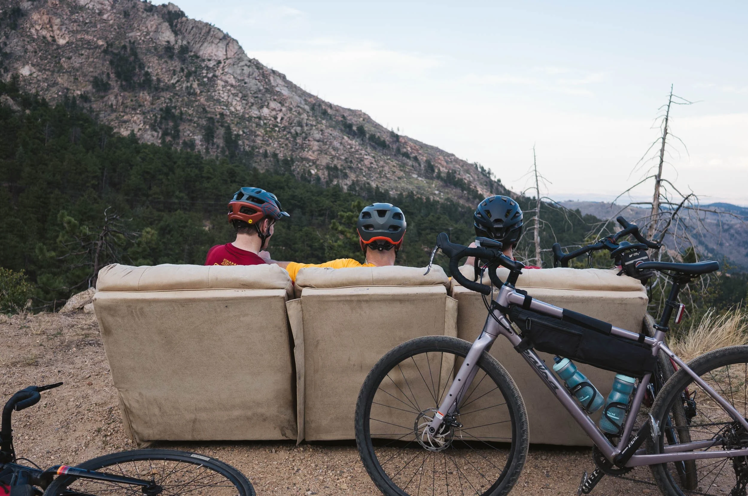 Three people with bicycle helmets sitting on a beige couch outdoors in a mountainous area, with bicycles parked nearby.