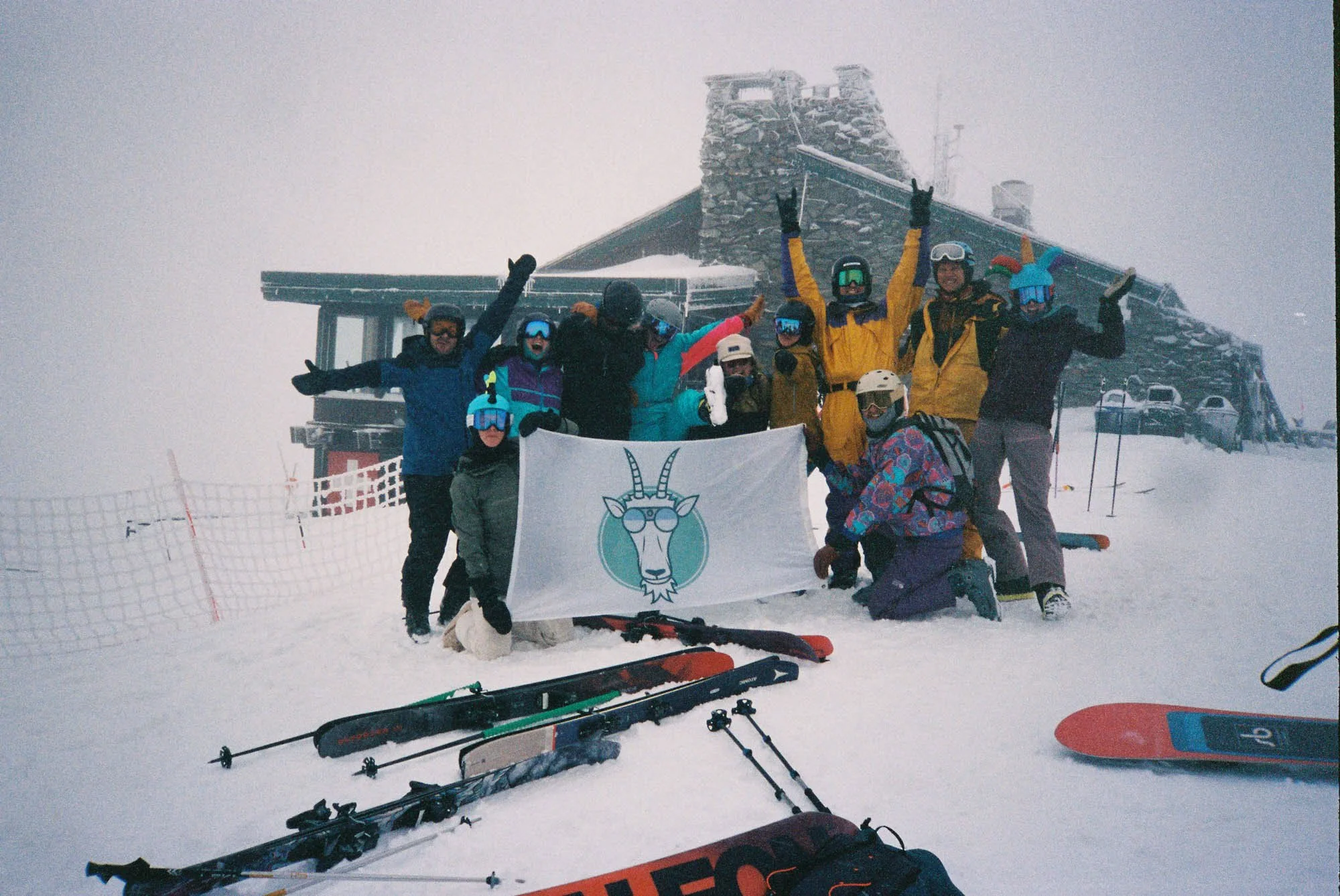 Group of skiers in colorful winter gear celebrating with raised arms in front of a snow-covered ski lodge during a snowstorm, with skis and ski poles on the snow in the foreground.