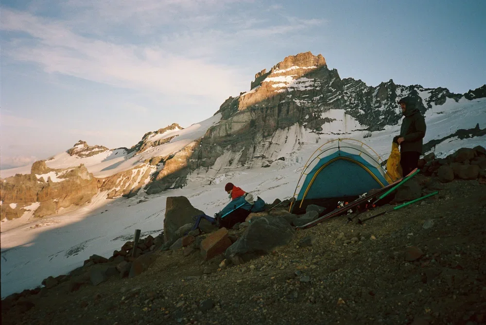 Two people set up a blue camping tent on rocky terrain in a mountainous area with snow and ice, with a mountain peak in the background during sunset or sunrise.