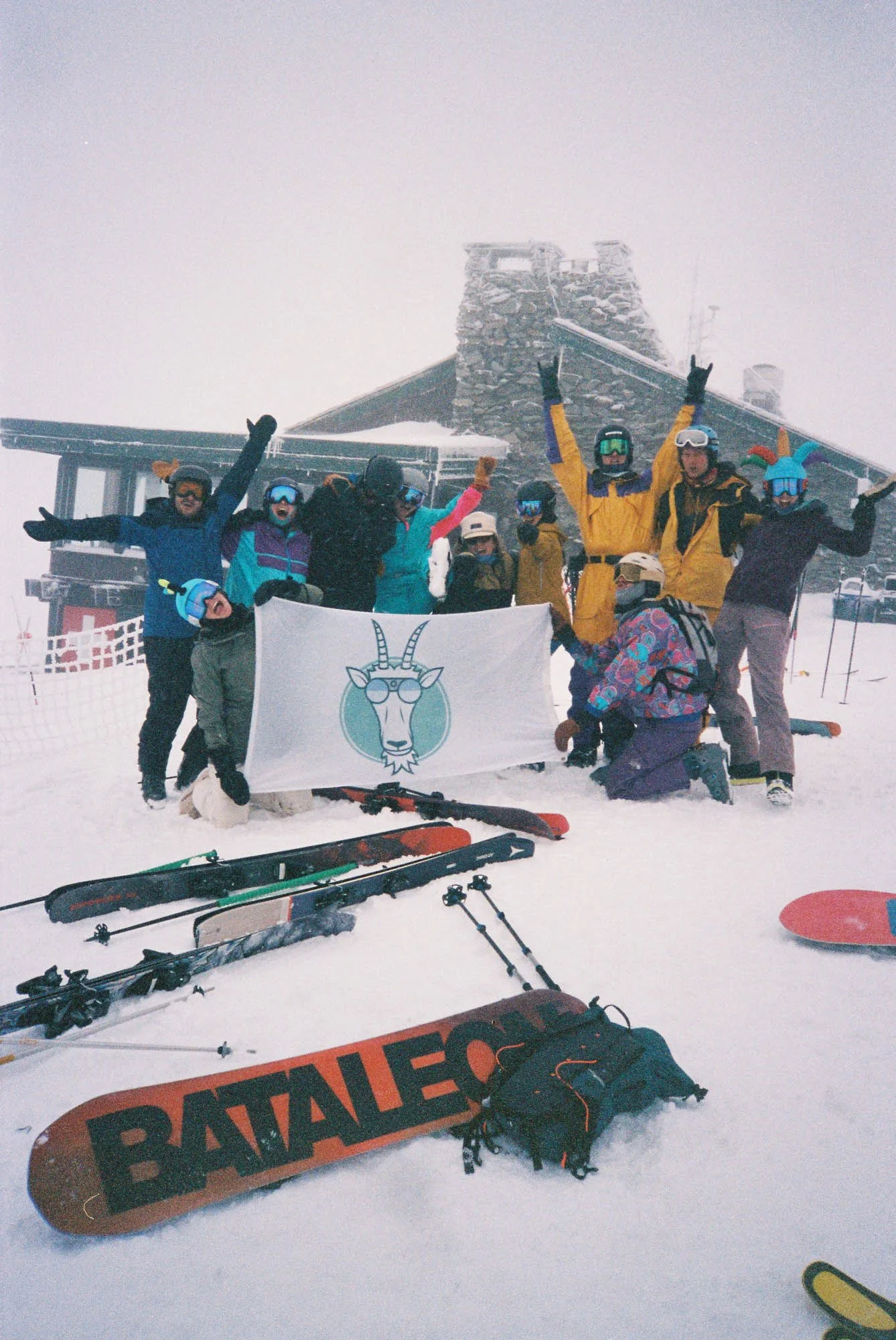 A group of skiers and snowboarders celebrating on a snowy mountain, holding a flag with a goat wearing sunglasses, in front of a wooden building with a tower and equipment. Ski gear and skis are scattered on the snow.