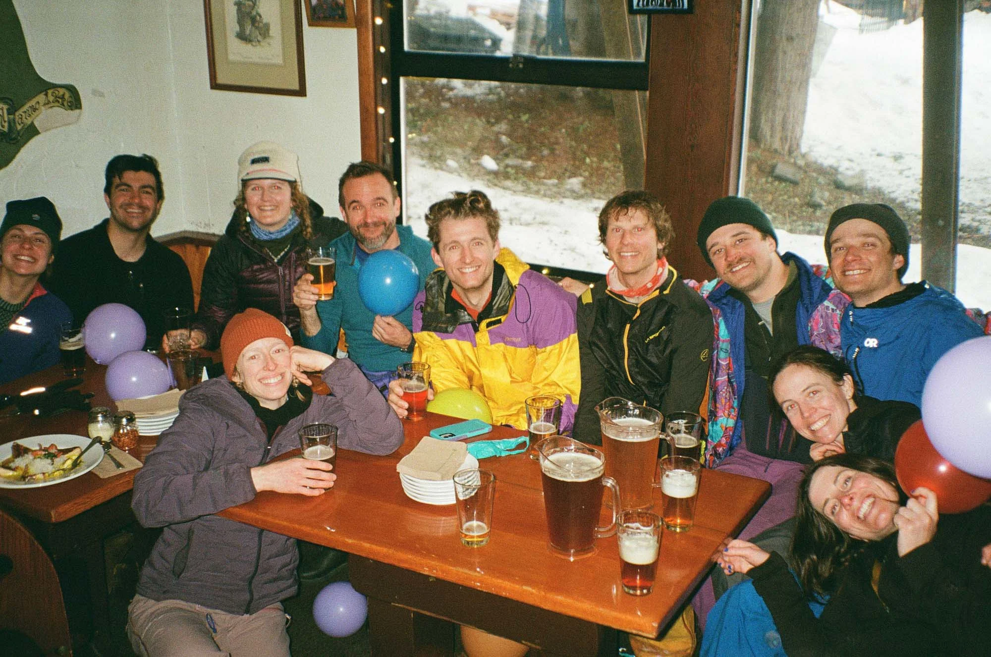 A group of people celebrating indoors at a gathering, sitting around a table with drinks and decorations, some smiling at the camera.