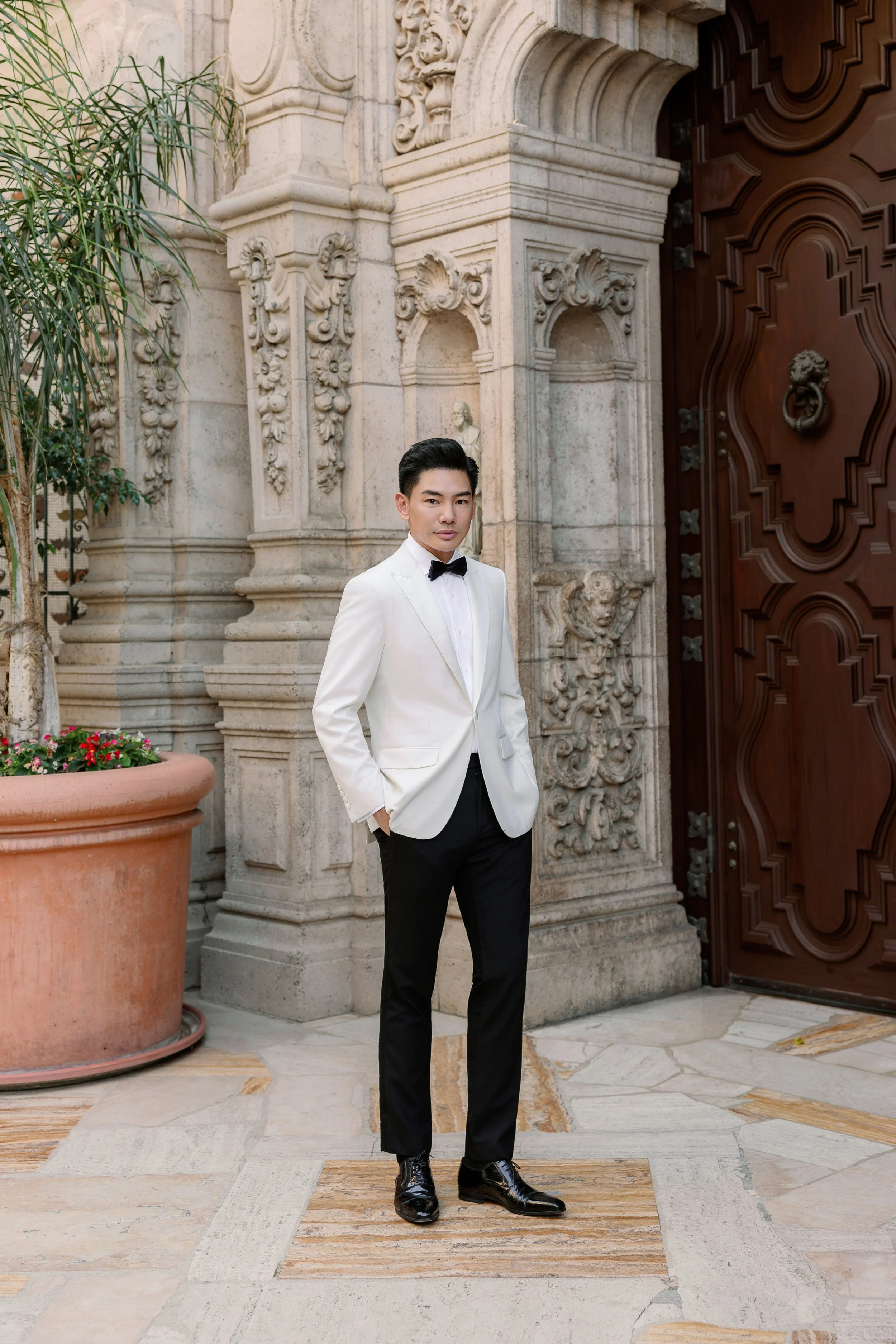 Young man in formal white tuxedo jacket, black bow tie, black pants, and shiny black shoes standing outside an ornate stone building with intricate carvings and a large wooden door.