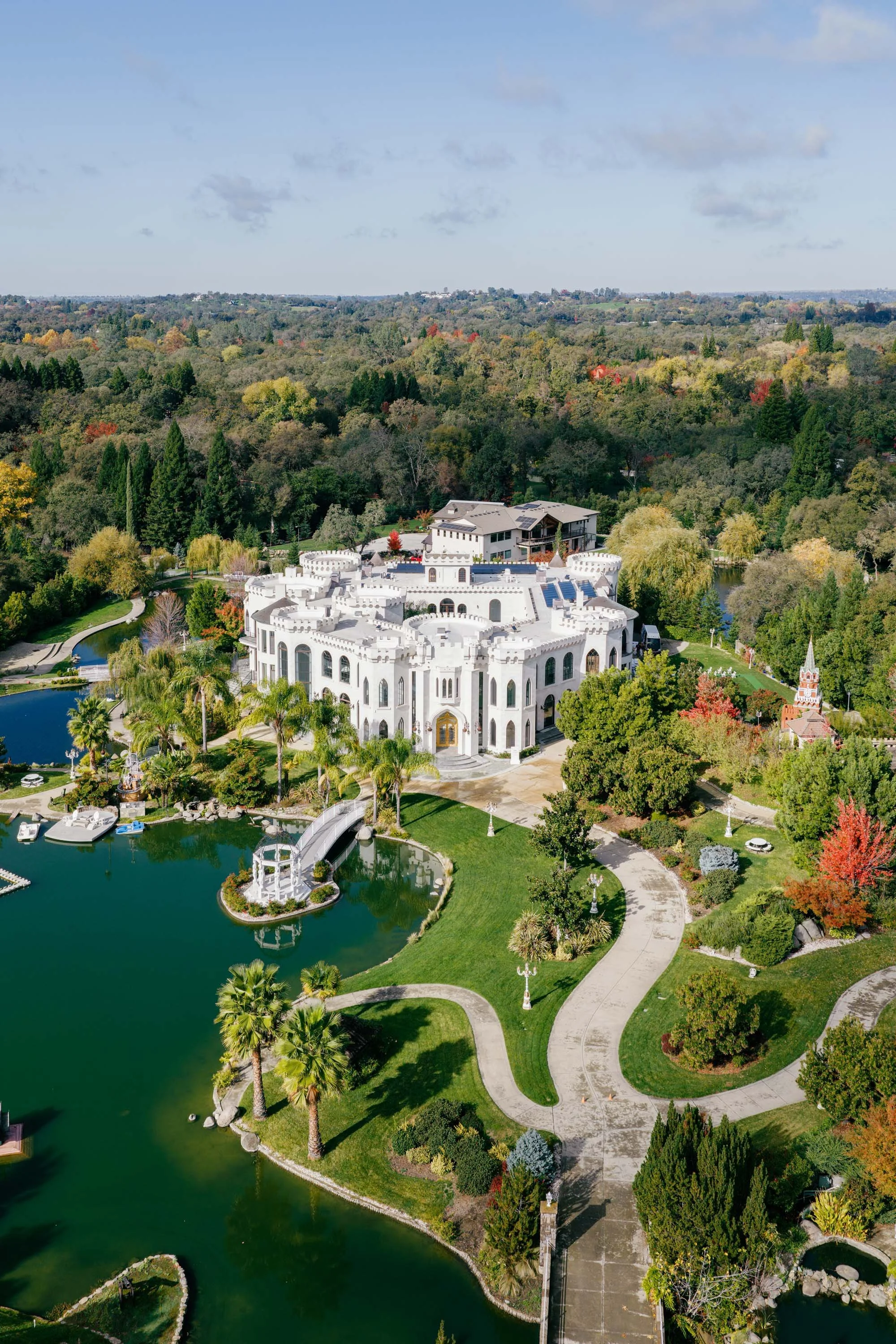 An aerial view of a large, castle-style mansion with white walls, surrounded by water and lush greenery, including trees with fall foliage.