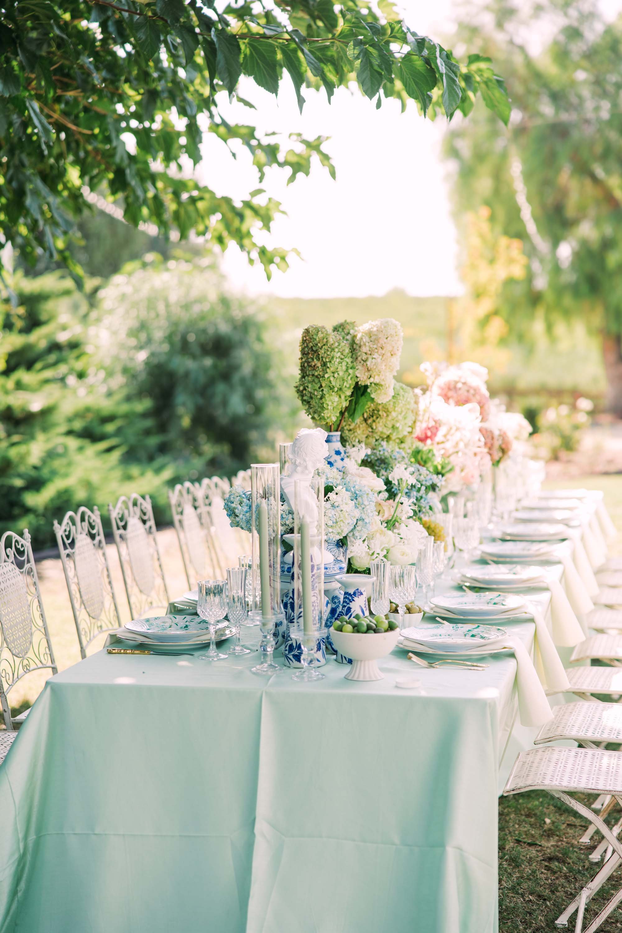 Outdoor table decorated with flowers, vases, and elegant place settings, set in a lush garden setting with sunlight filtering through trees.