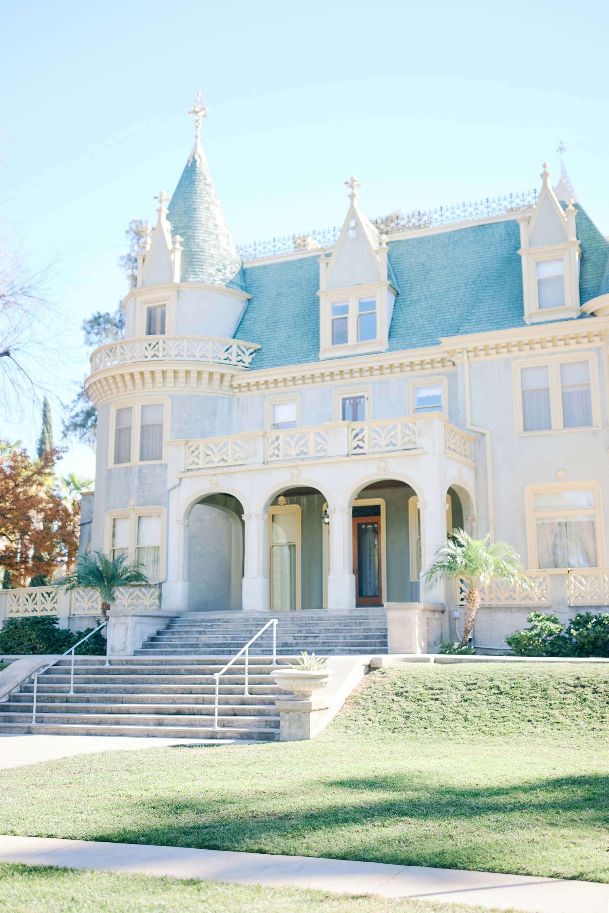 A large, ornate Victorian-style house with a blue roof, turret, and balcony, surrounded by a well-maintained lawn and tropical plants.