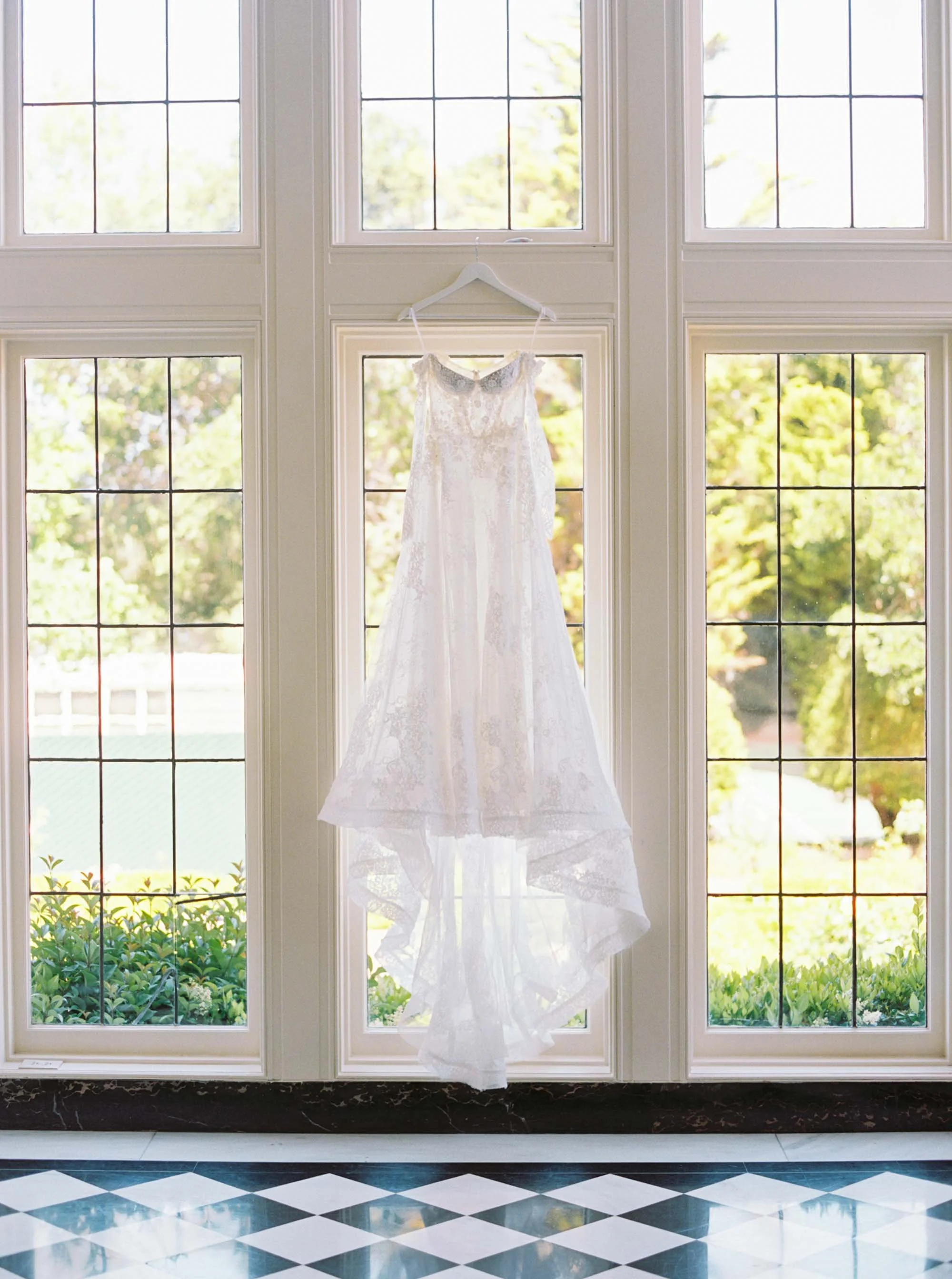 A white wedding dress hangs on a hanger in front of a large set of windows, with sunlight illuminating the dress. Green foliage is visible outside the windows, and the floor has a black and white checkered pattern.
