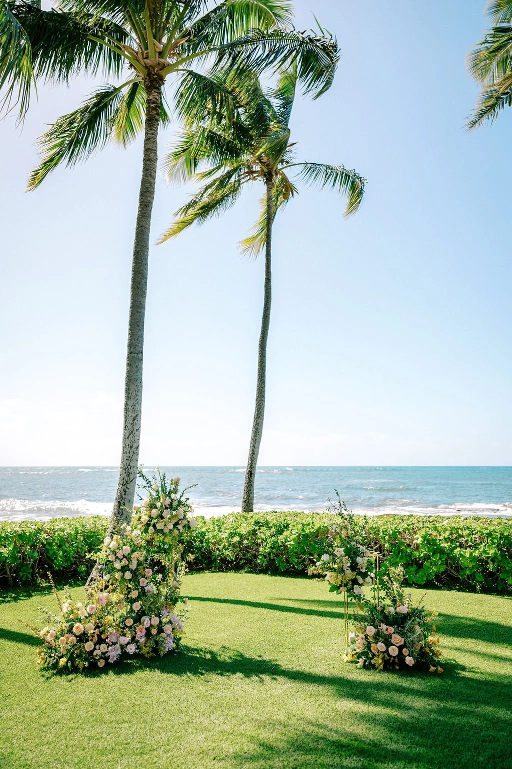 Beachside wedding setup with two palm trees, a grassy area, and floral arrangements at the base of the trees, overlooking the ocean with a clear sky.