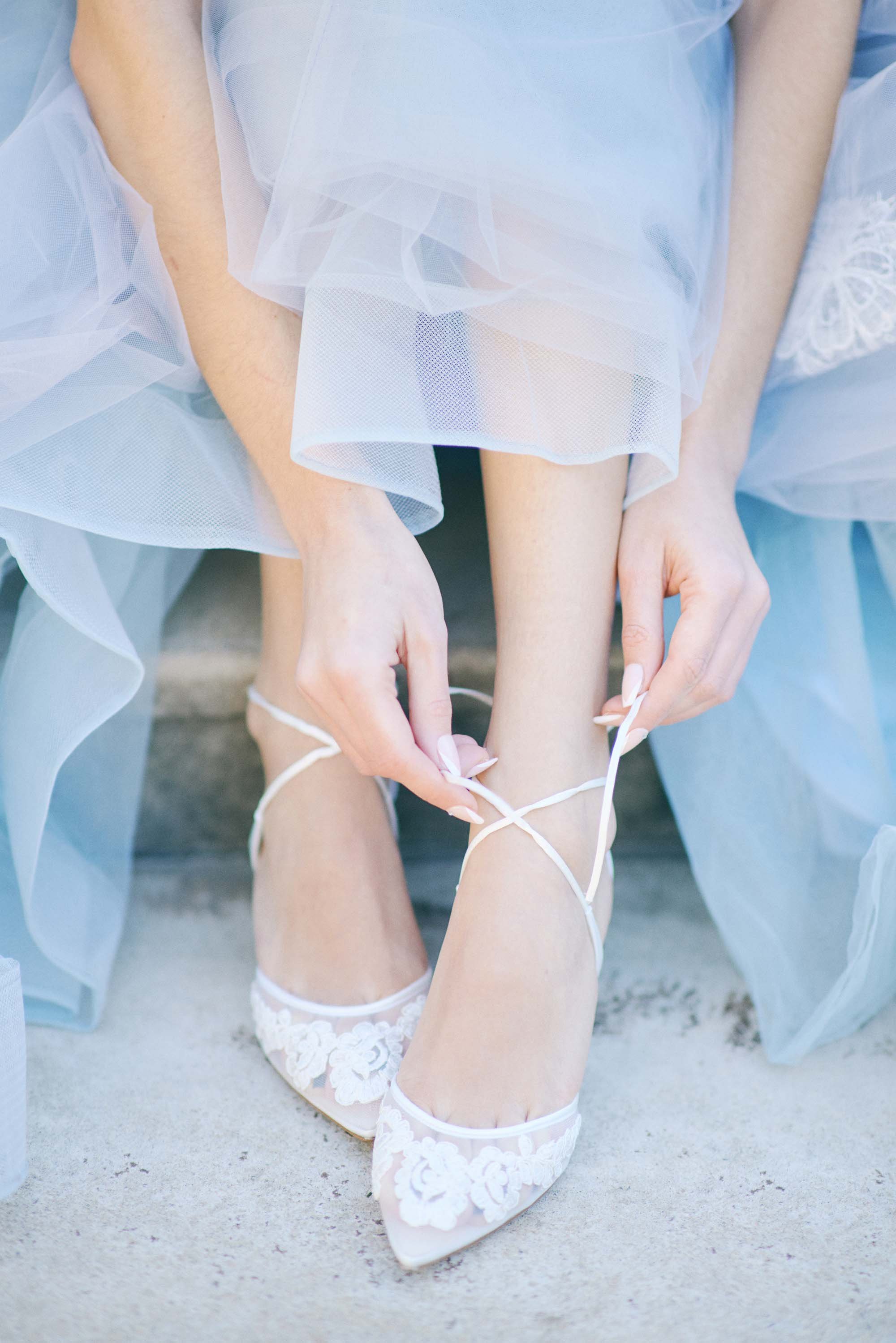 A woman in a light blue or white dress is tying the laces of her white, lace detailed high-heeled shoes. The dress appears to be made of sheer, layered fabric.