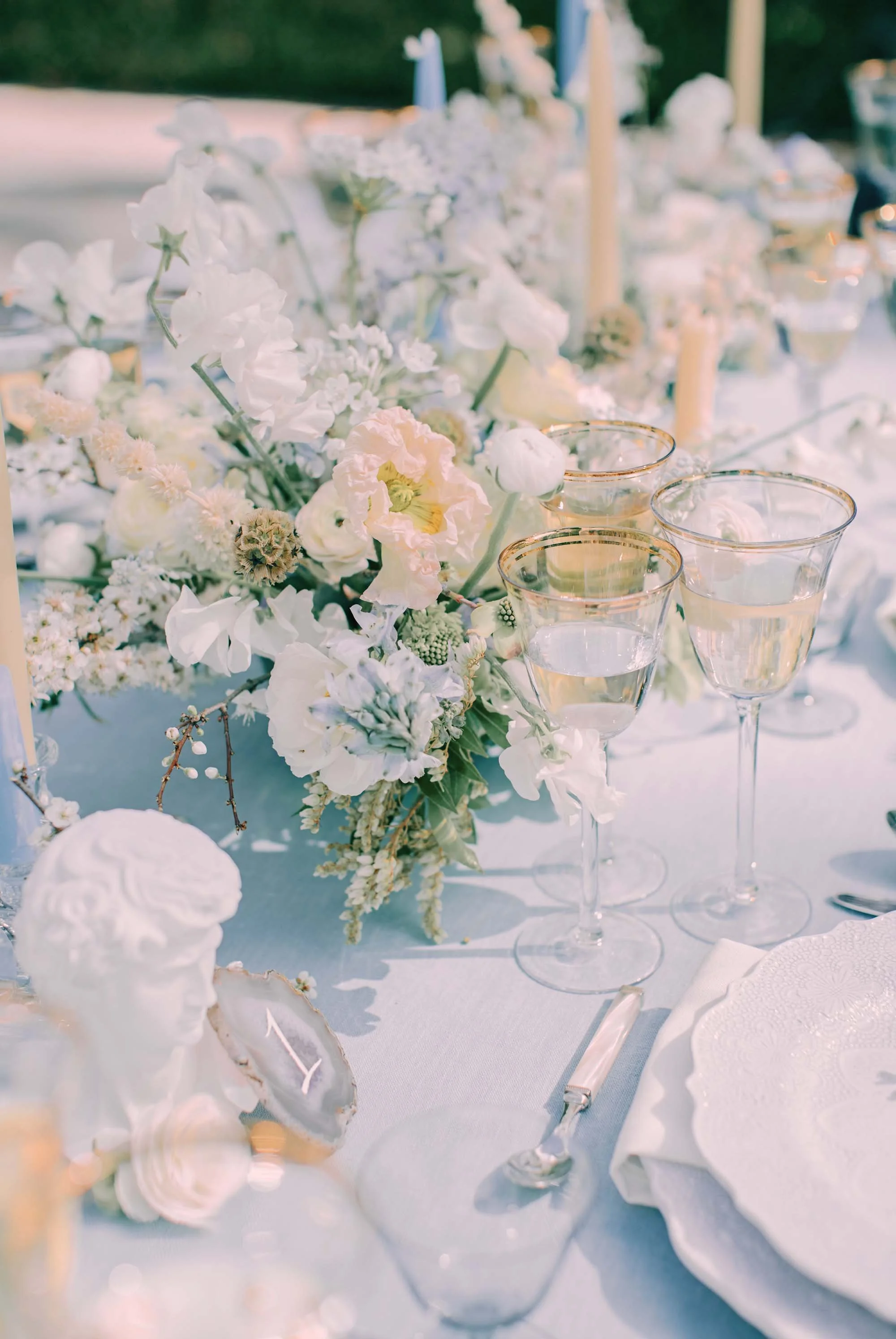 Elegant outdoor table setting with a floral centerpiece, white and pastel flowers, wine glasses filled with white wine, a white plate with a scalloped edge, a knife, and white cloth napkin.