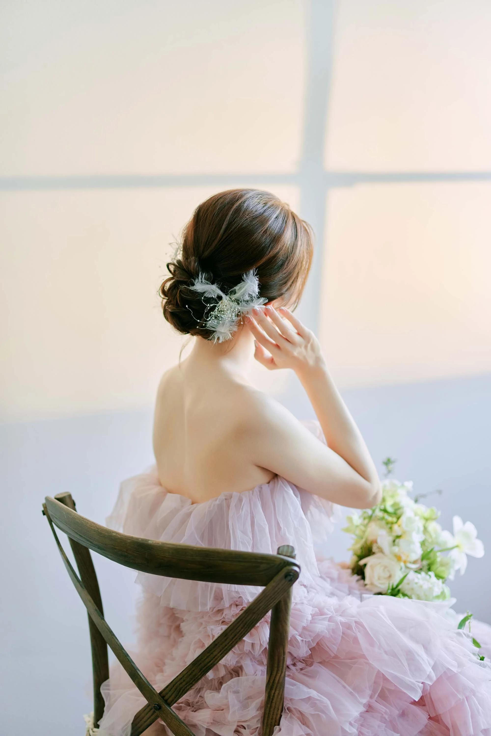 A woman with a floral hair accessory sitting on a wooden chair in a light pink, ruffled dress, with a bouquet of white flowers nearby, looking away from the camera.