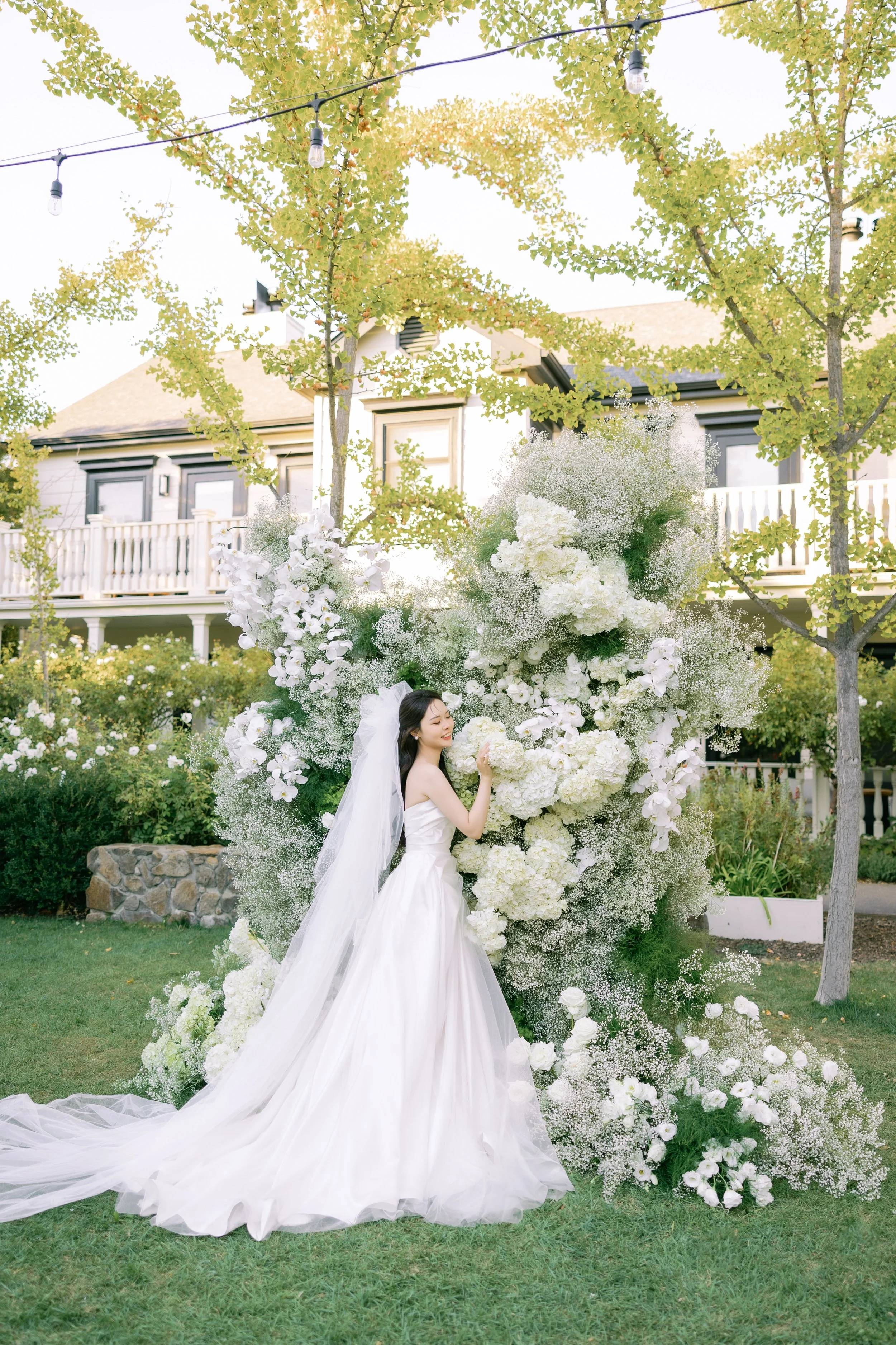 A woman in traditional Korean wedding hanbok standing outdoors surrounded by white and pink flowers with trees in the background.