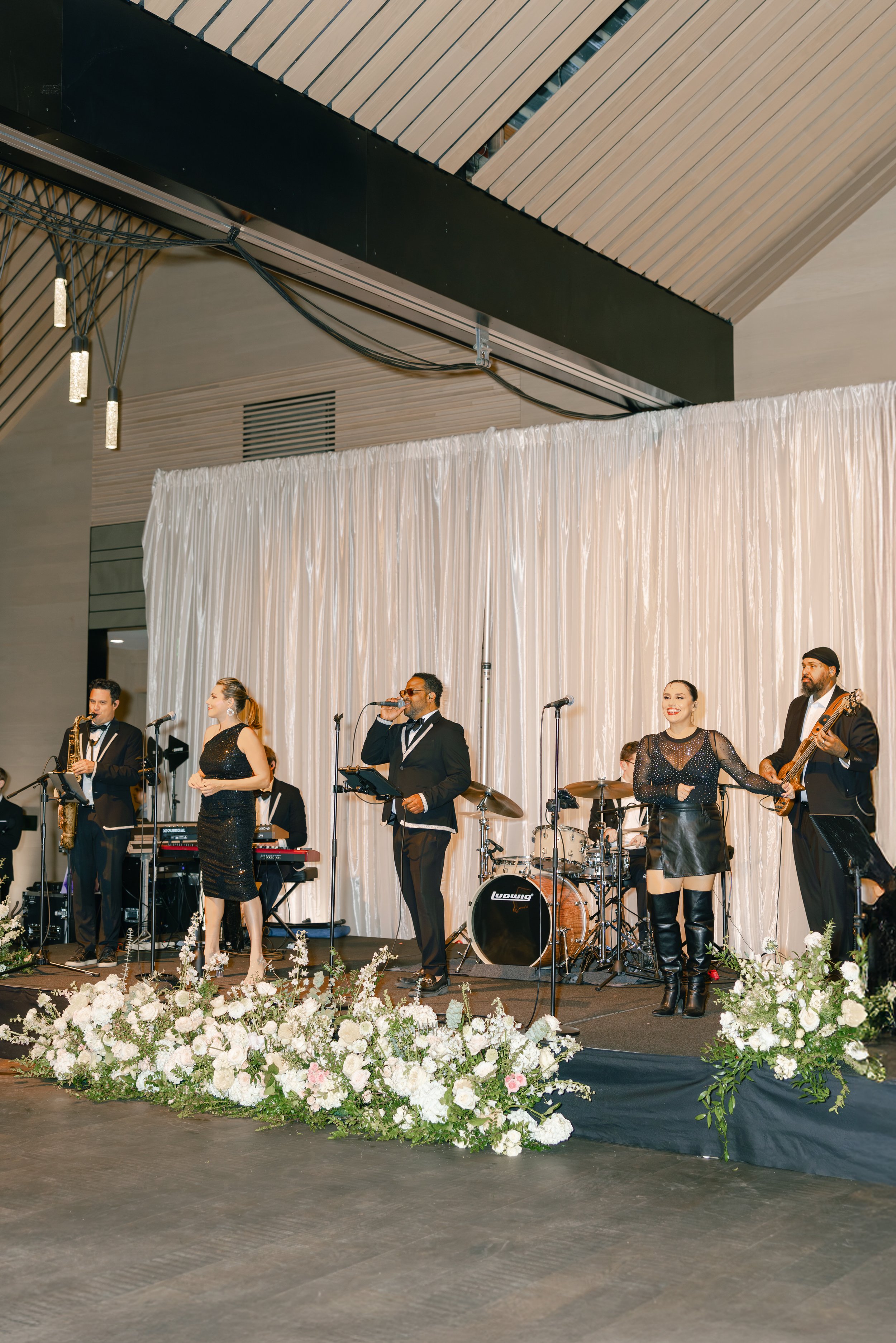 A live band performing on stage in a decorated venue with white floral arrangements in front.