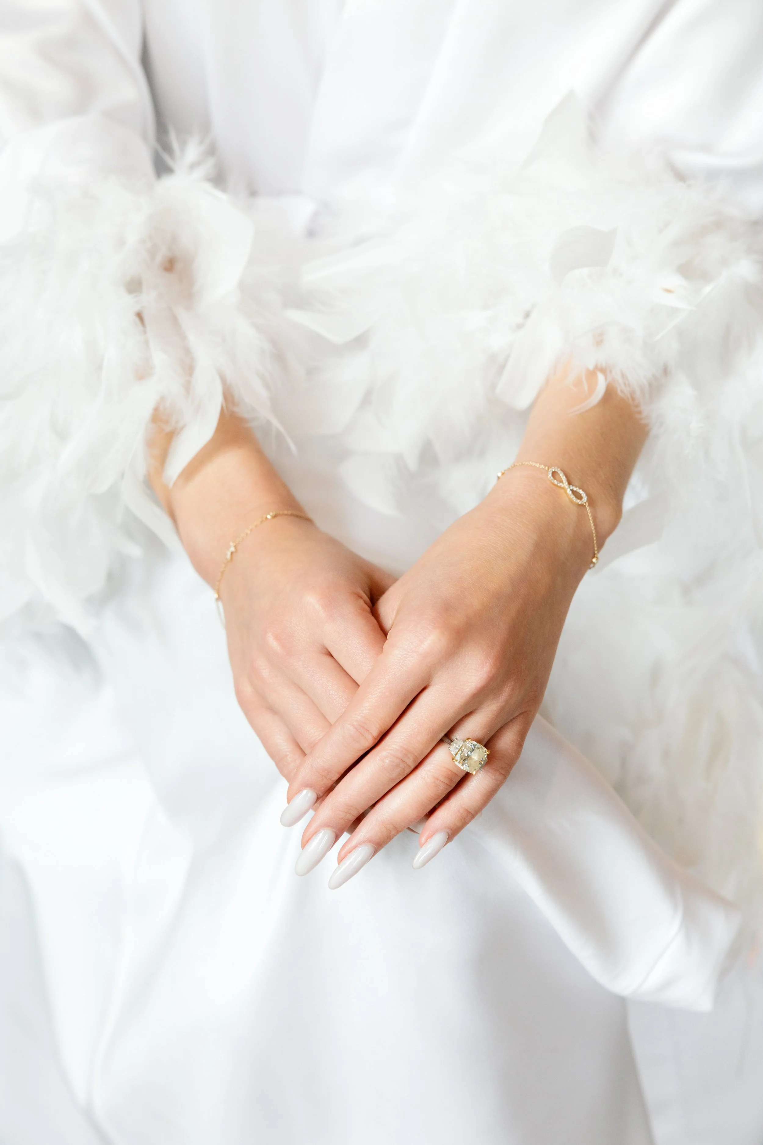 Close-up of a woman's hands with manicured nails and jewelry, resting on a white fabric background adorned with white feathers.