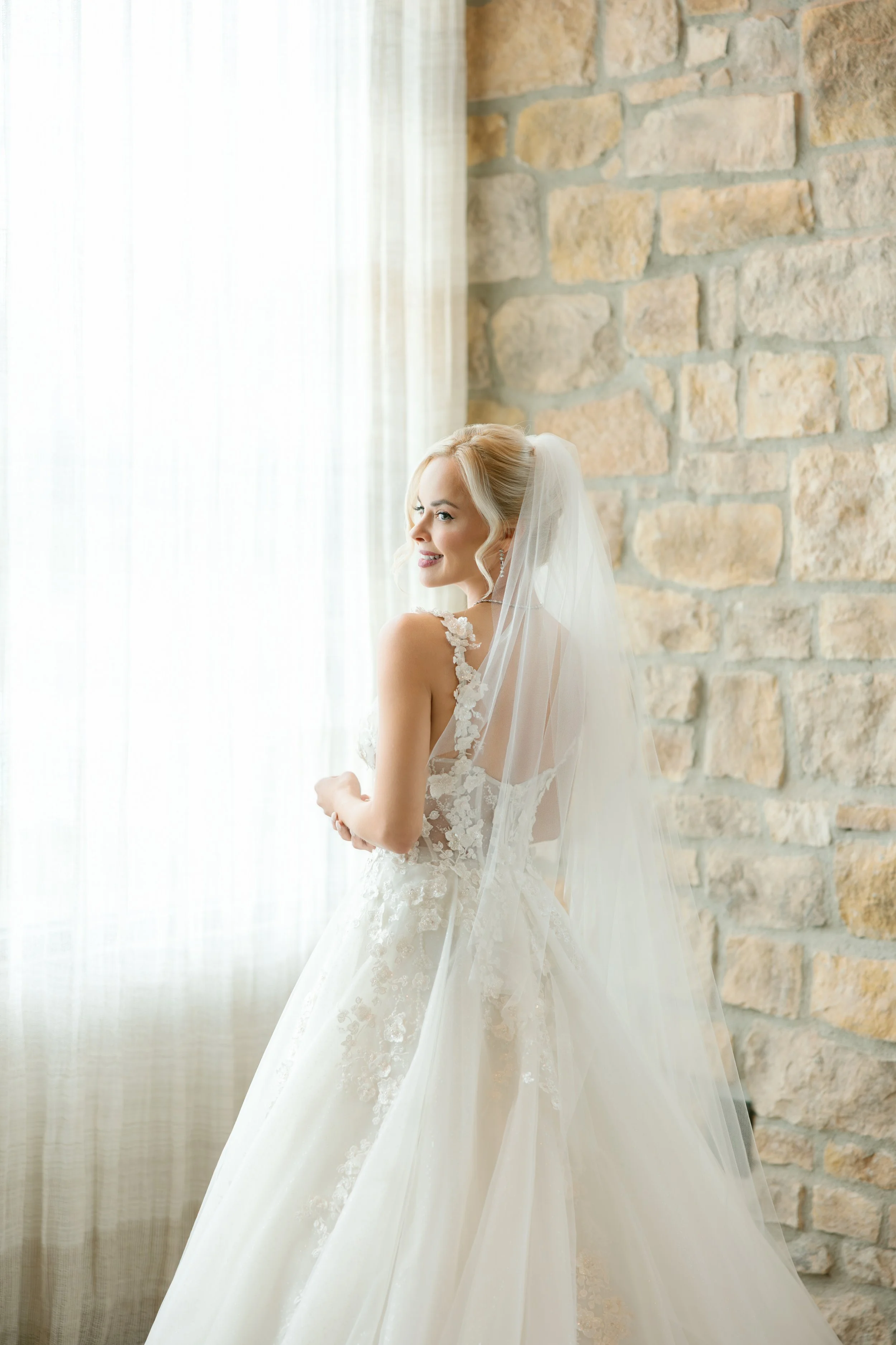 A bride in a wedding dress with lace details, standing by a window with sheer curtains, smiling and looking outside, with a stone wall in the background.
