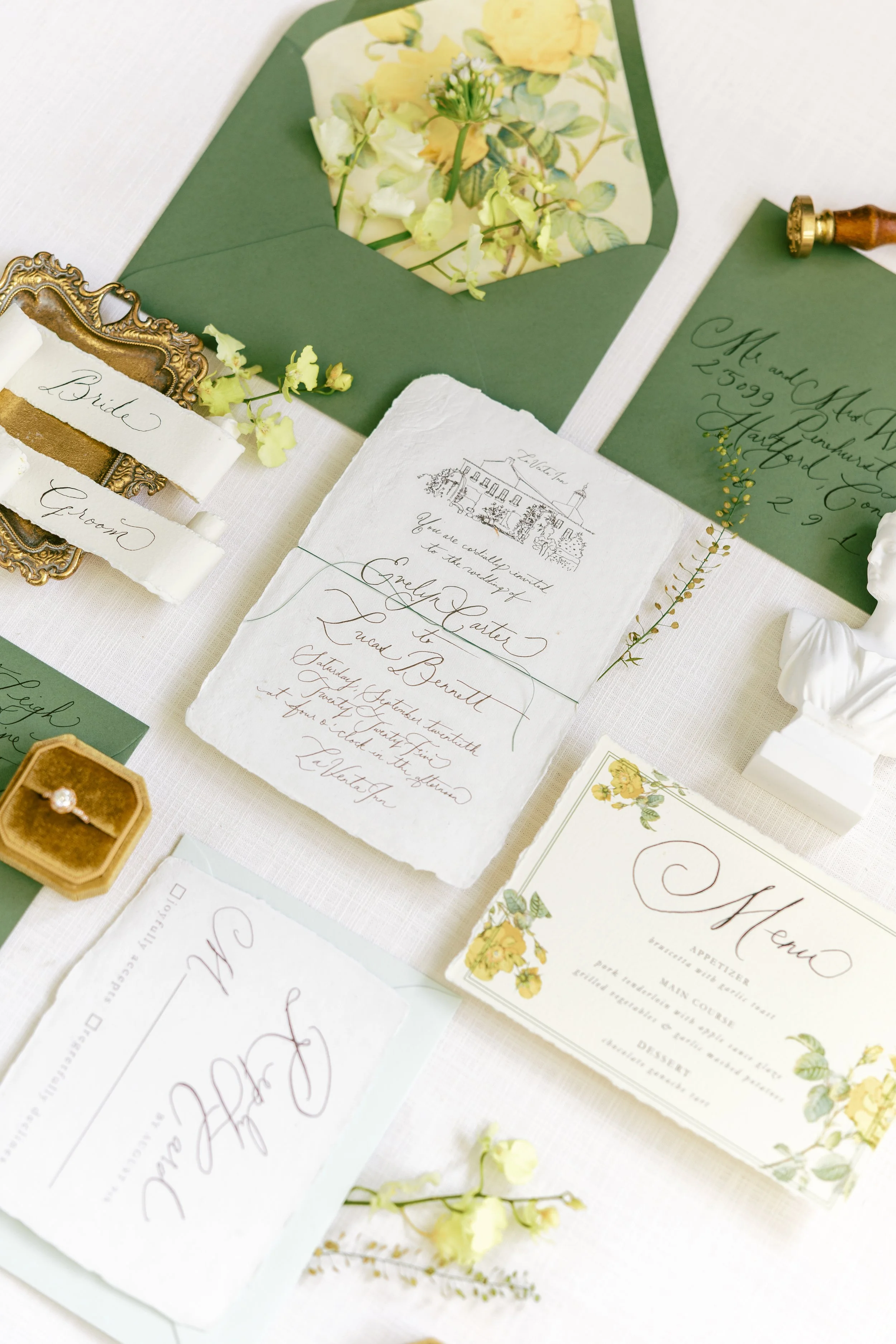 Wedding invitation suite with menus, RSVP card, address envelope, and small decorative flowers on a white tablecloth.