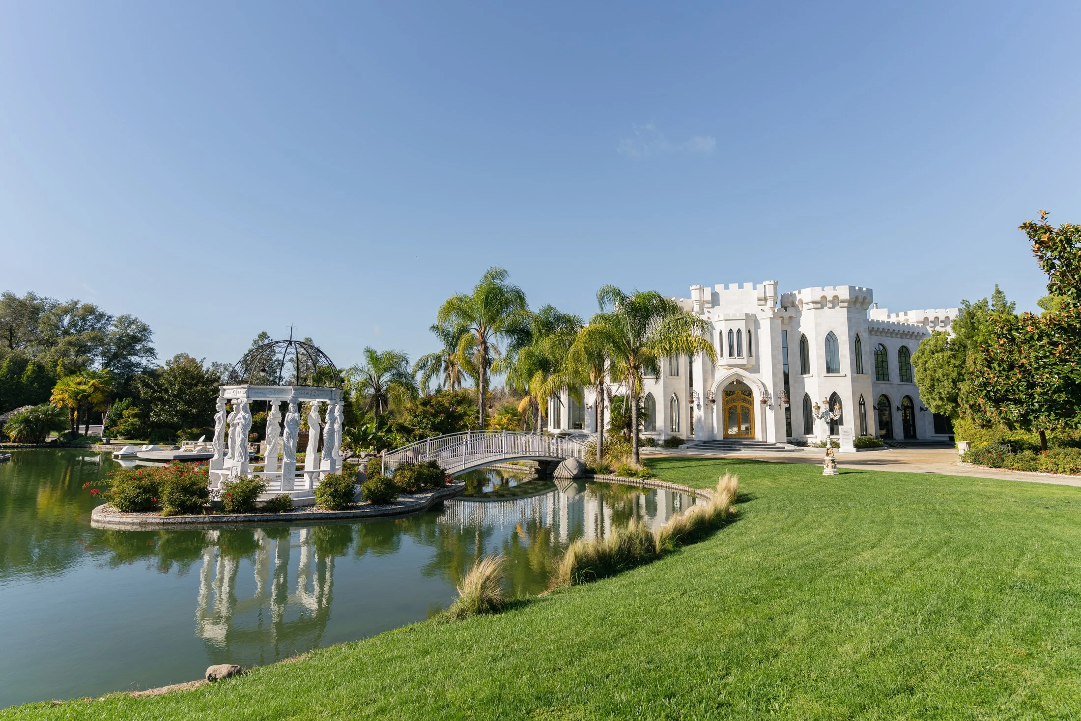 Exterior view of White Swan Castle wedding venue with a lake, bridge, palm trees, and manicured lawn.