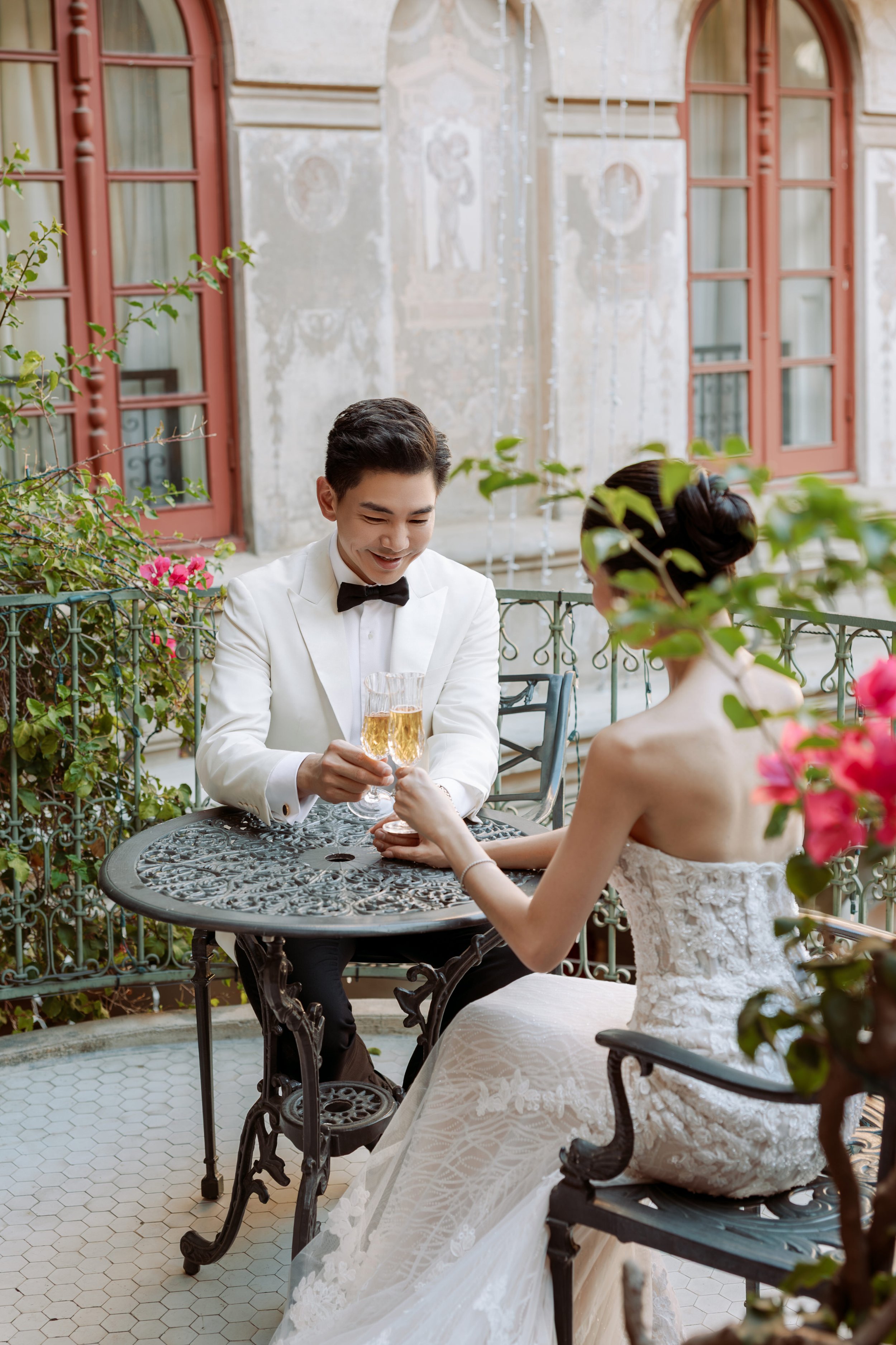 A bride and groom sitting at a small outdoor table on a balcony, toasting with champagne glasses, surrounded by flowers and greenery.