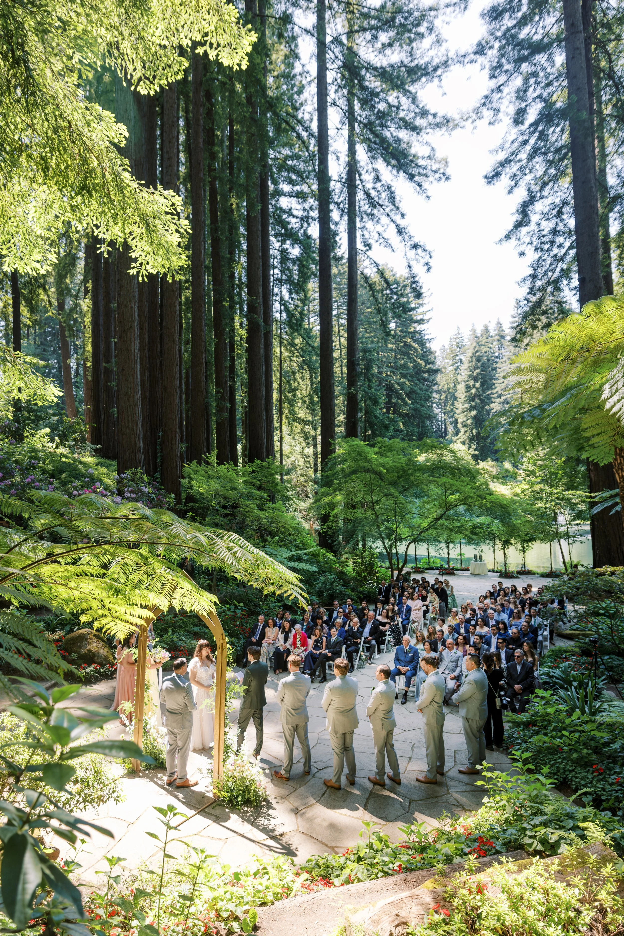 Outdoor wedding ceremony taking place in a lush forest with tall trees and vibrant green foliage. The bride and groom are standing under a wooden arch, surrounded by an audience seated on chairs.