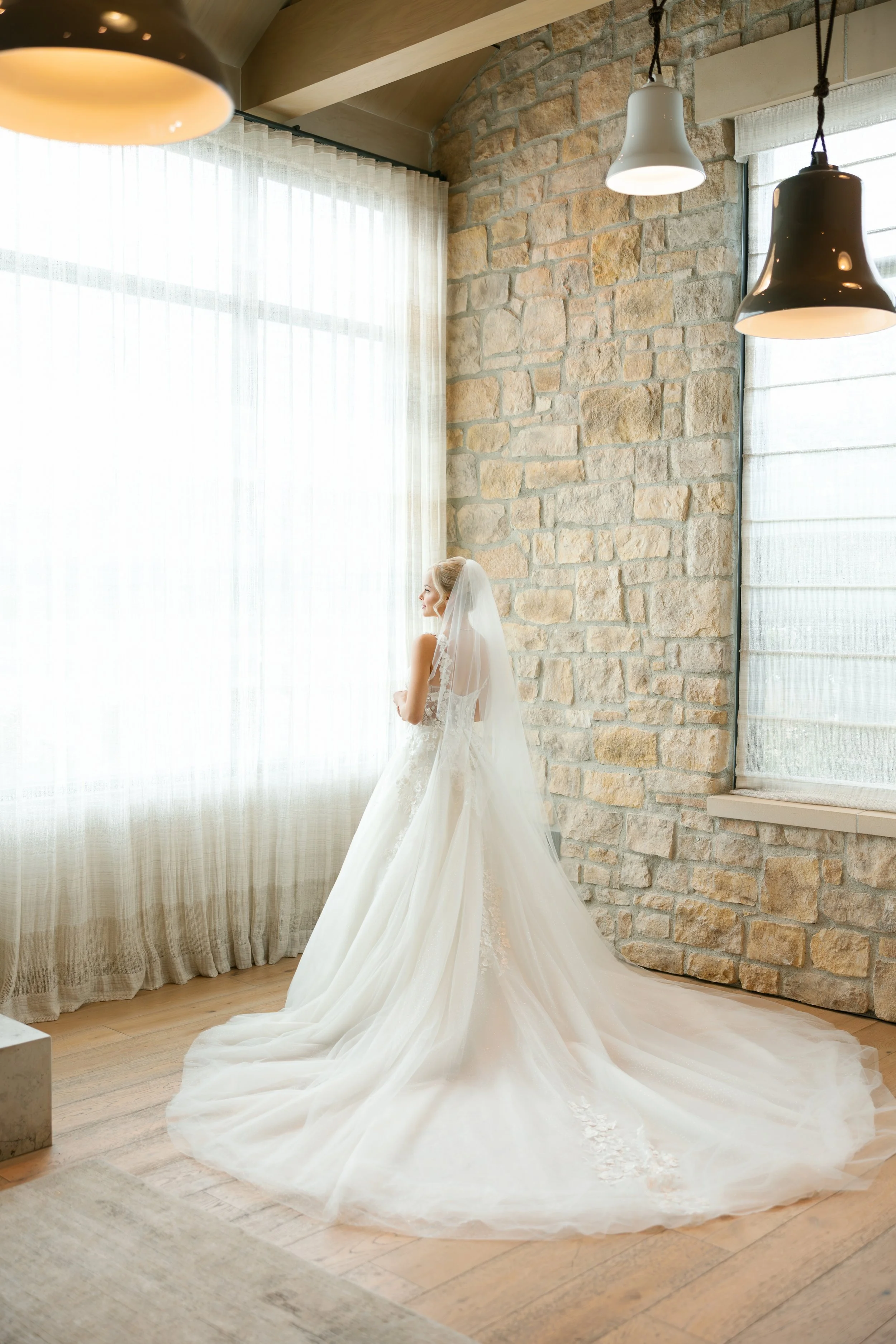 Bride wearing a wedding dress and veil standing by a large window with sheer curtains, in a room with stone walls and hanging light fixtures.