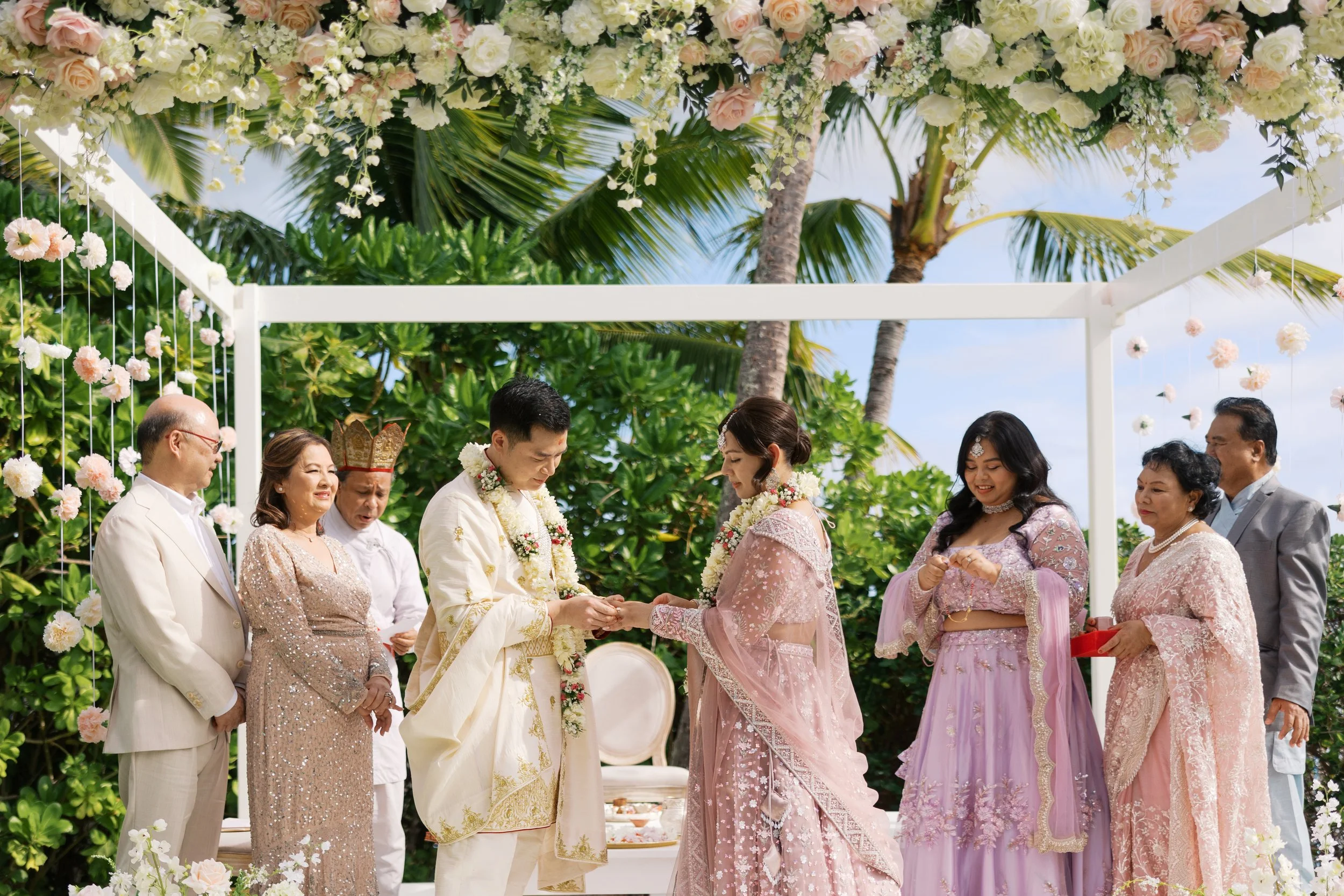 A traditional wedding ceremony taking place outdoors under a floral arch with tropical palm trees in the background. The bride and groom are exchanging rings, surrounded by family members dressed in colorful, elegant attire.