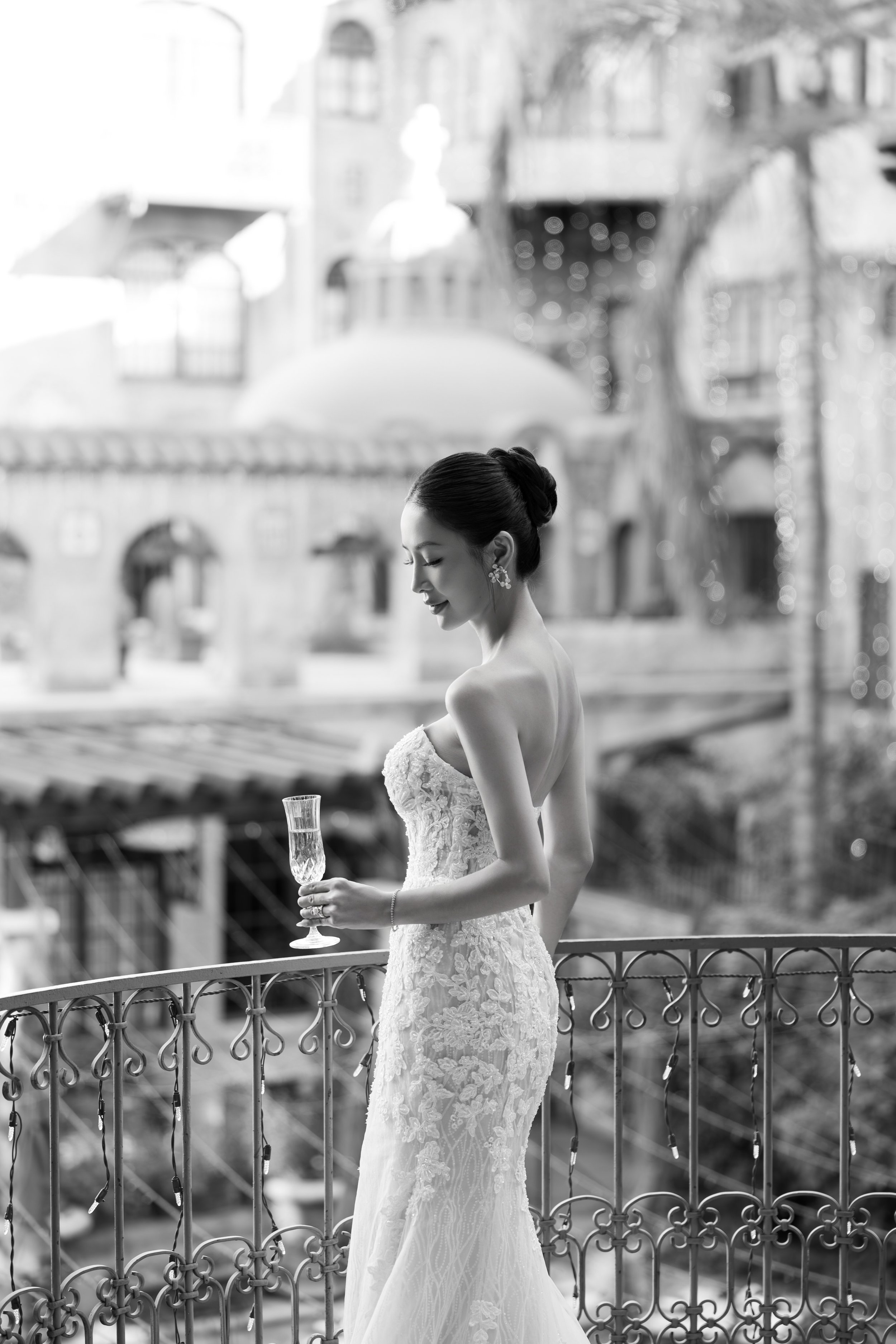 A woman in a wedding dress holding a glass of champagne, standing on a balcony with an ornate railing, in front of blurred European-style buildings and lights.