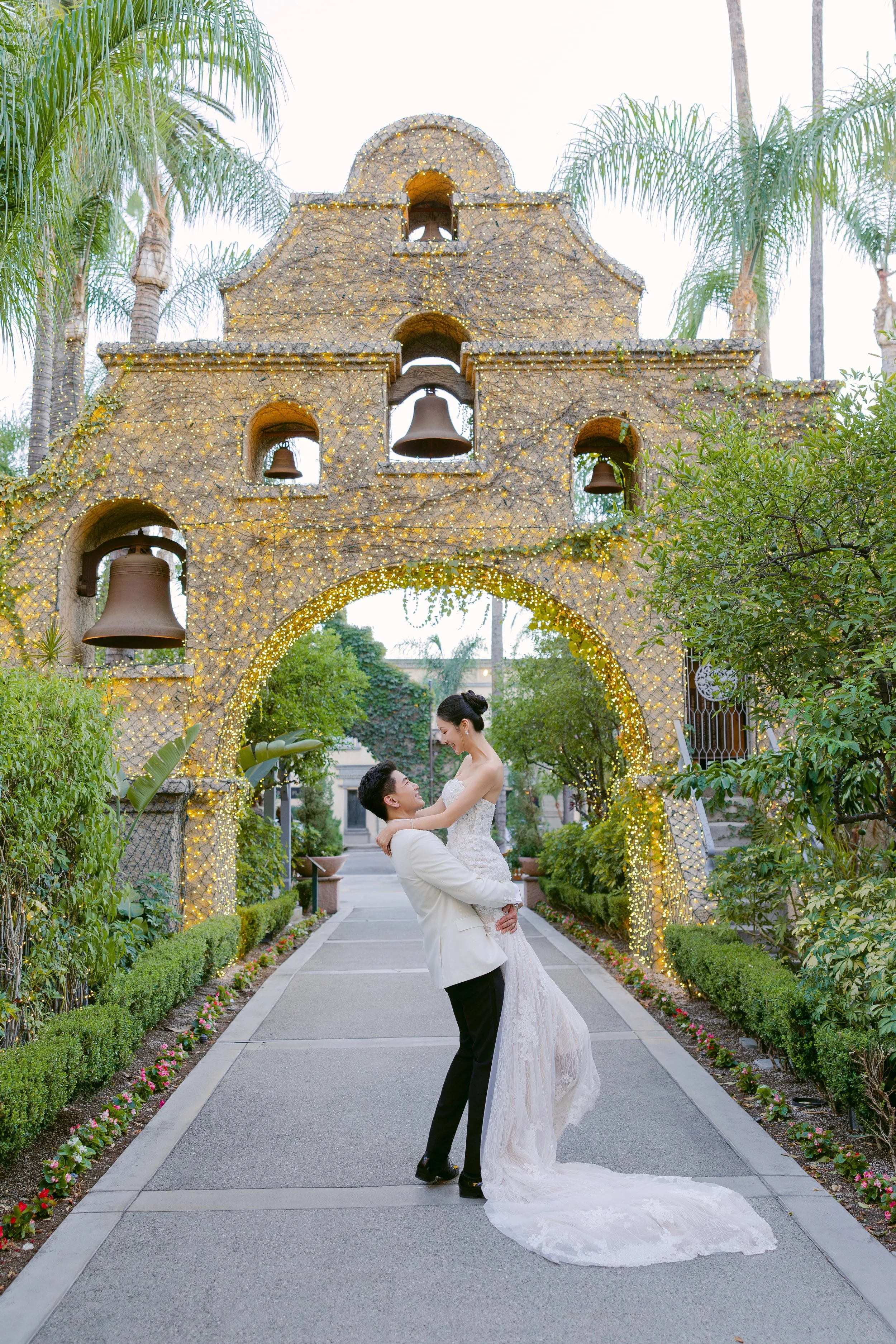 A wedding couple with the groom lifting the bride under a decorative archway with bells and fairy lights in a garden setting.