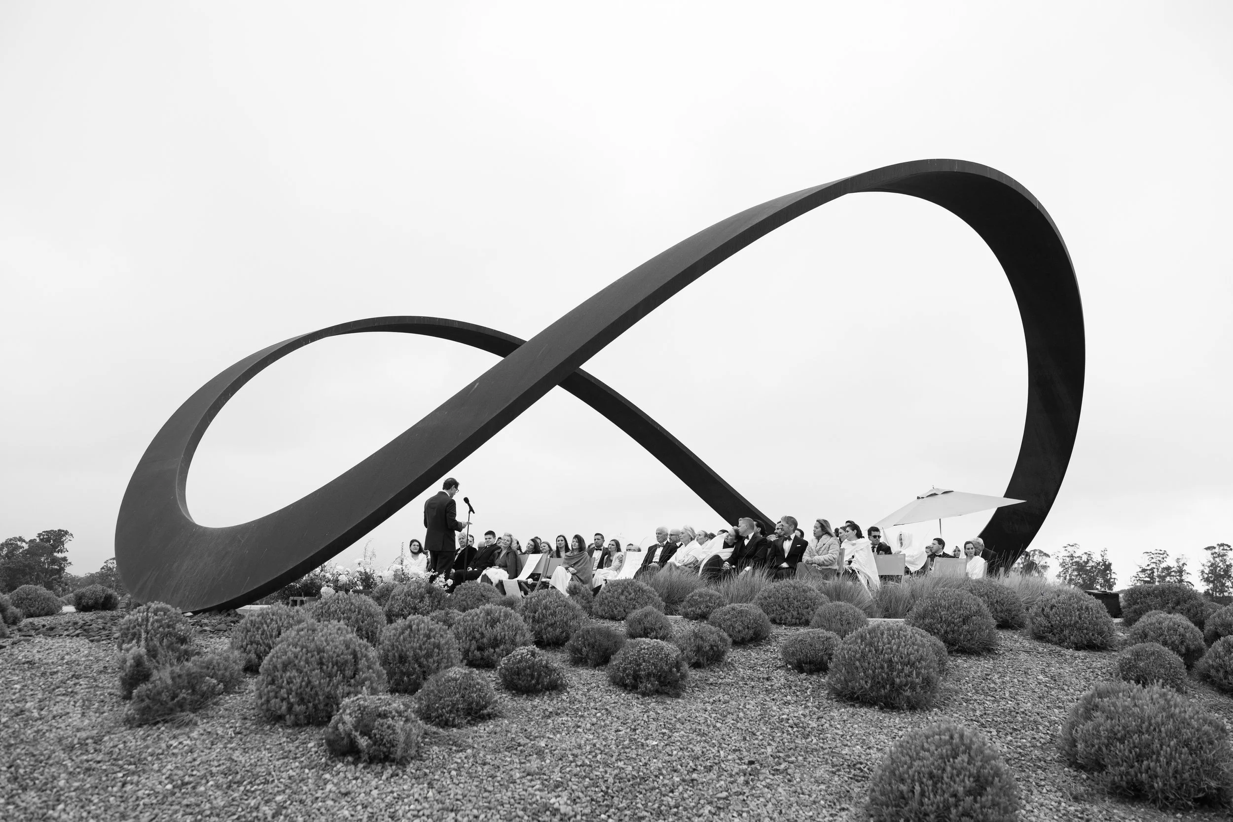 A group of people attending an outdoor event or ceremony, seated on chairs, with a speaker standing in front of them, under a large, abstract, looped metal sculpture on a landscaped area with bushes. The image is in black and white.