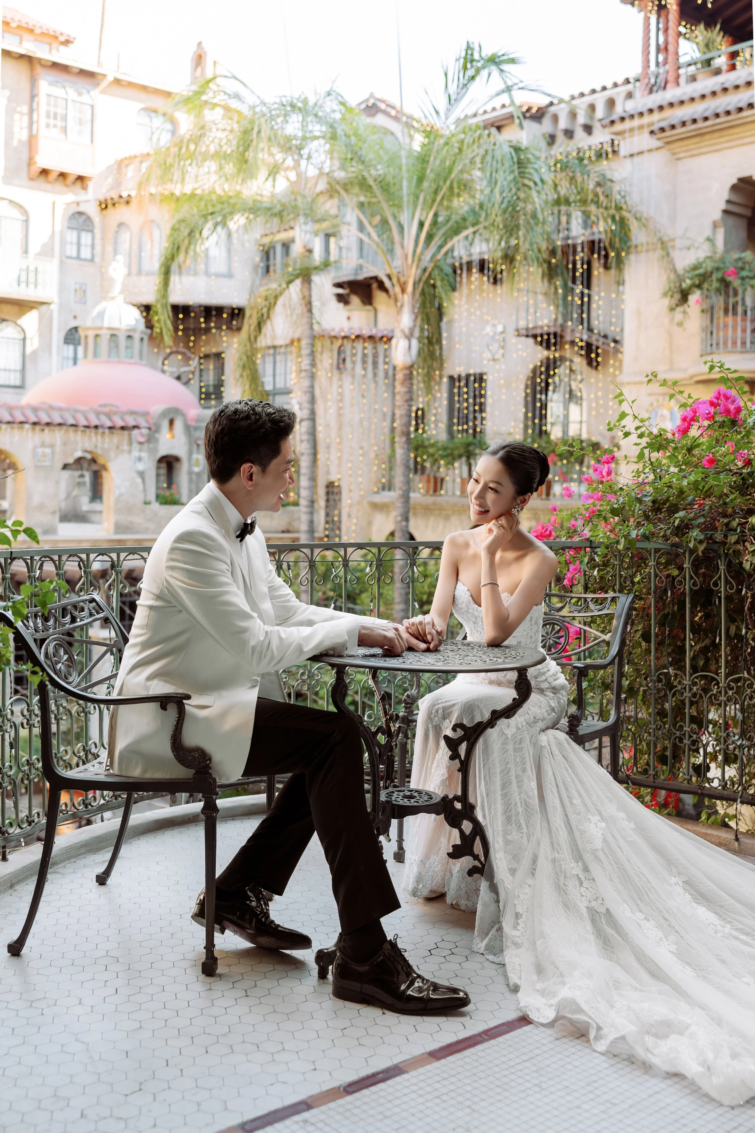 A newlywed couple in wedding attire sitting at a small table on a balcony with flowers, palm trees, and colorful buildings in the background, enjoying a moment together.