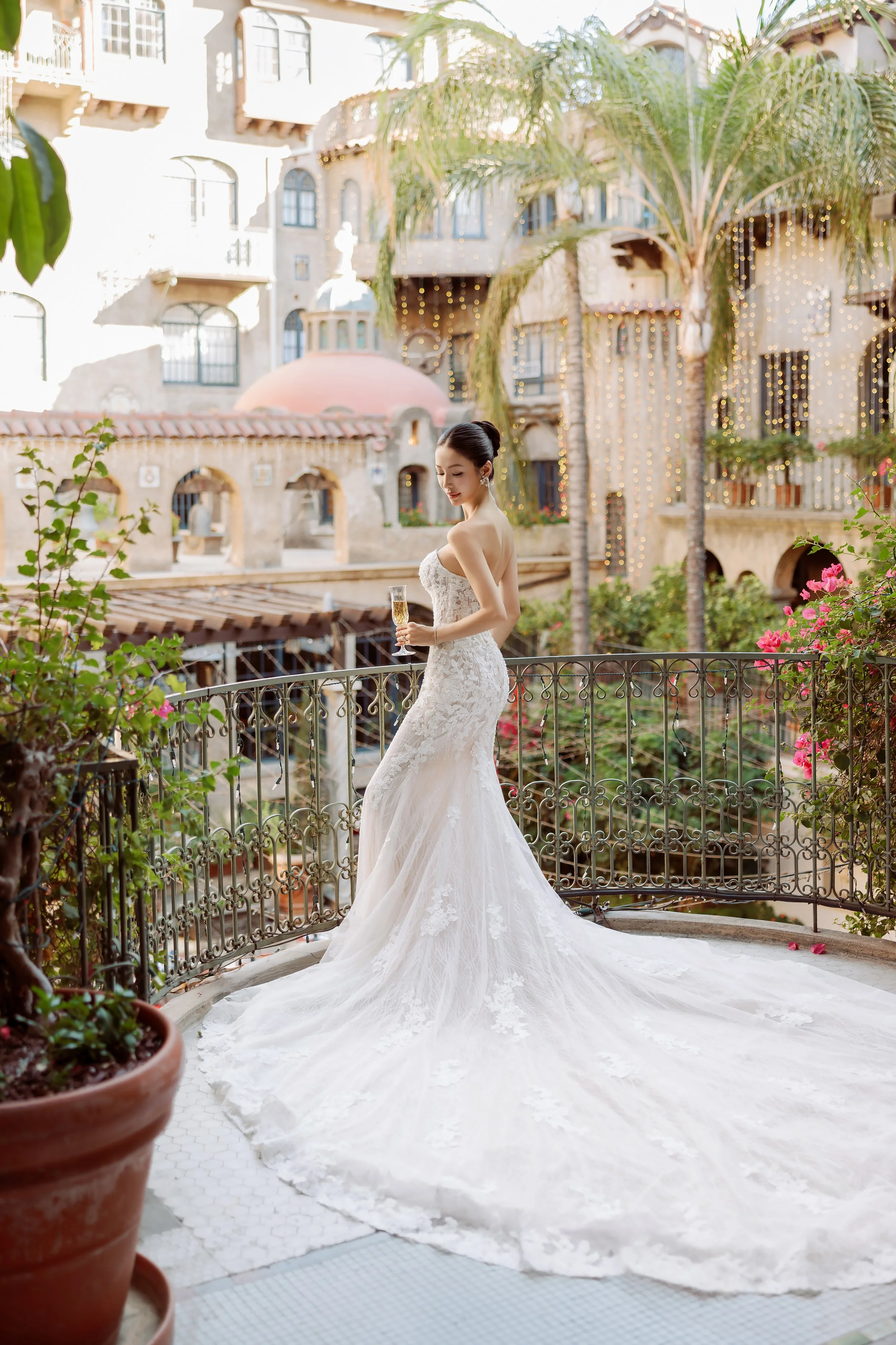 A bride in a strapless white lace wedding gown holding a glass of champagne on a balcony with cityscape and string lights behind her.