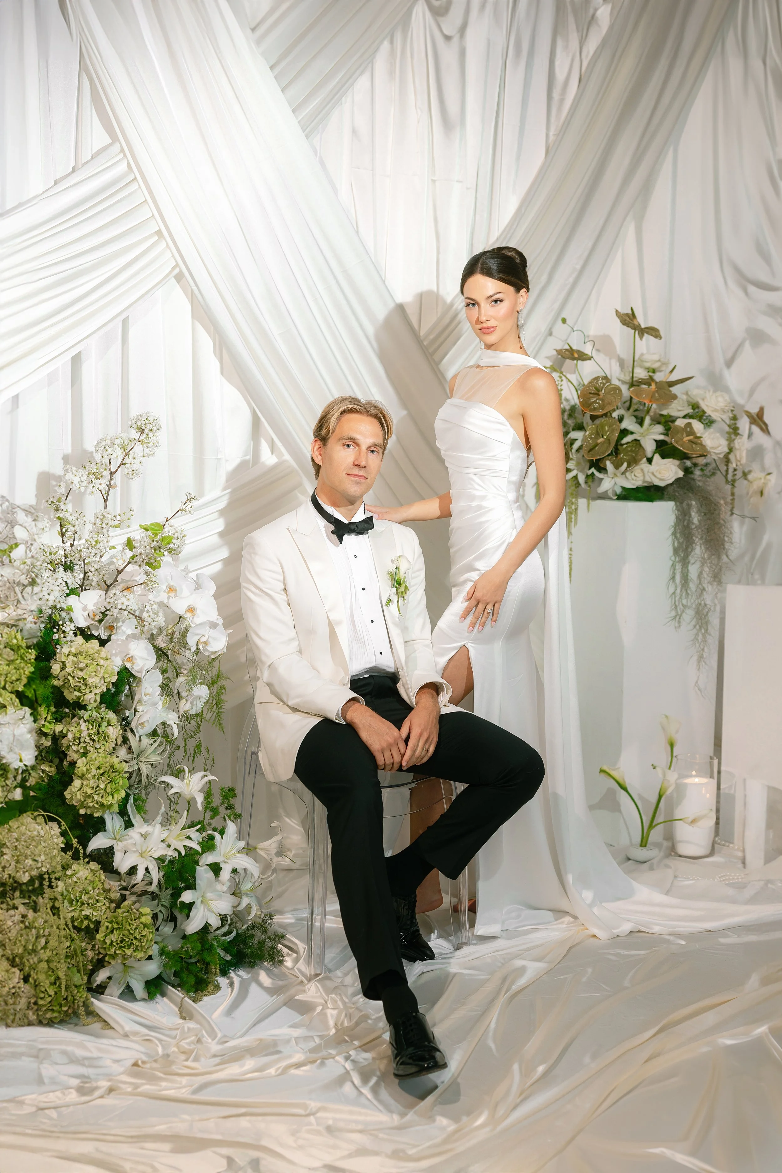A woman in a light gray wedding dress holding a bouquet of white and pastel flowers outdoors with green trees in the background.