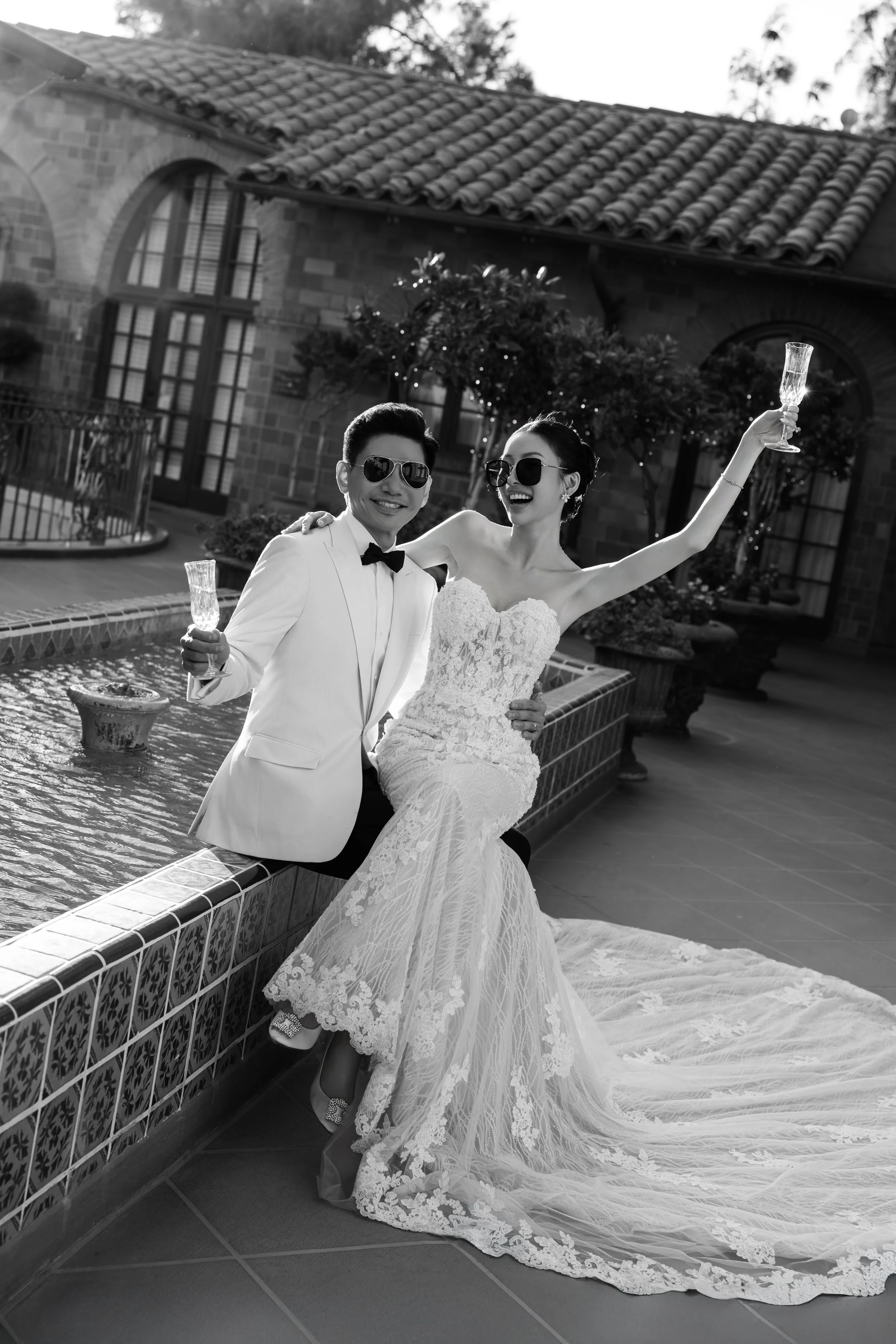 A black and white photo of a bride and groom celebrating outdoors, sitting on the edge of a fountain, both wearing sunglasses and holding champagne glasses, with a building and greenery in the background.