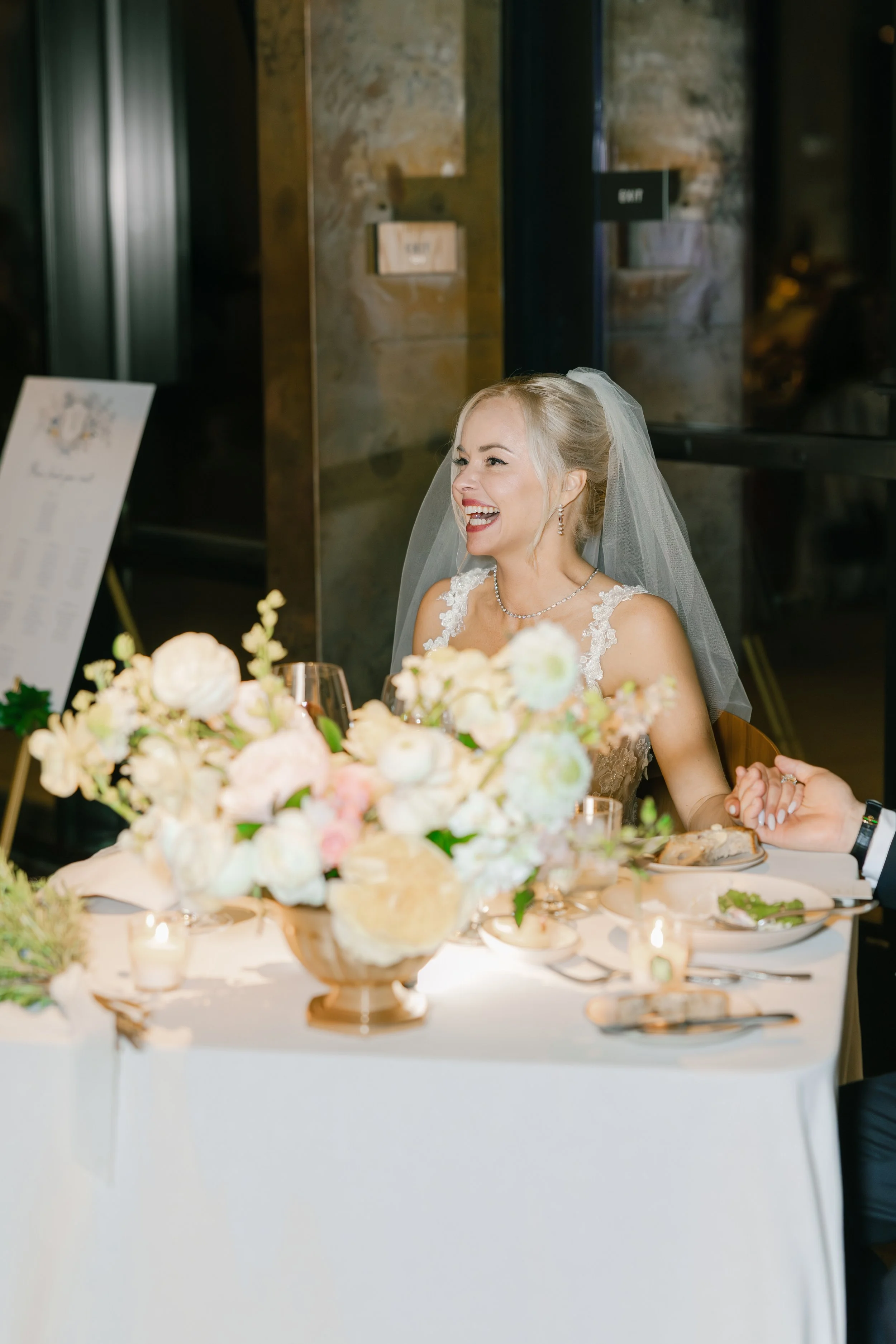 Bride with blonde hair and veil, smiling, sitting at a wedding reception table with floral centerpiece.