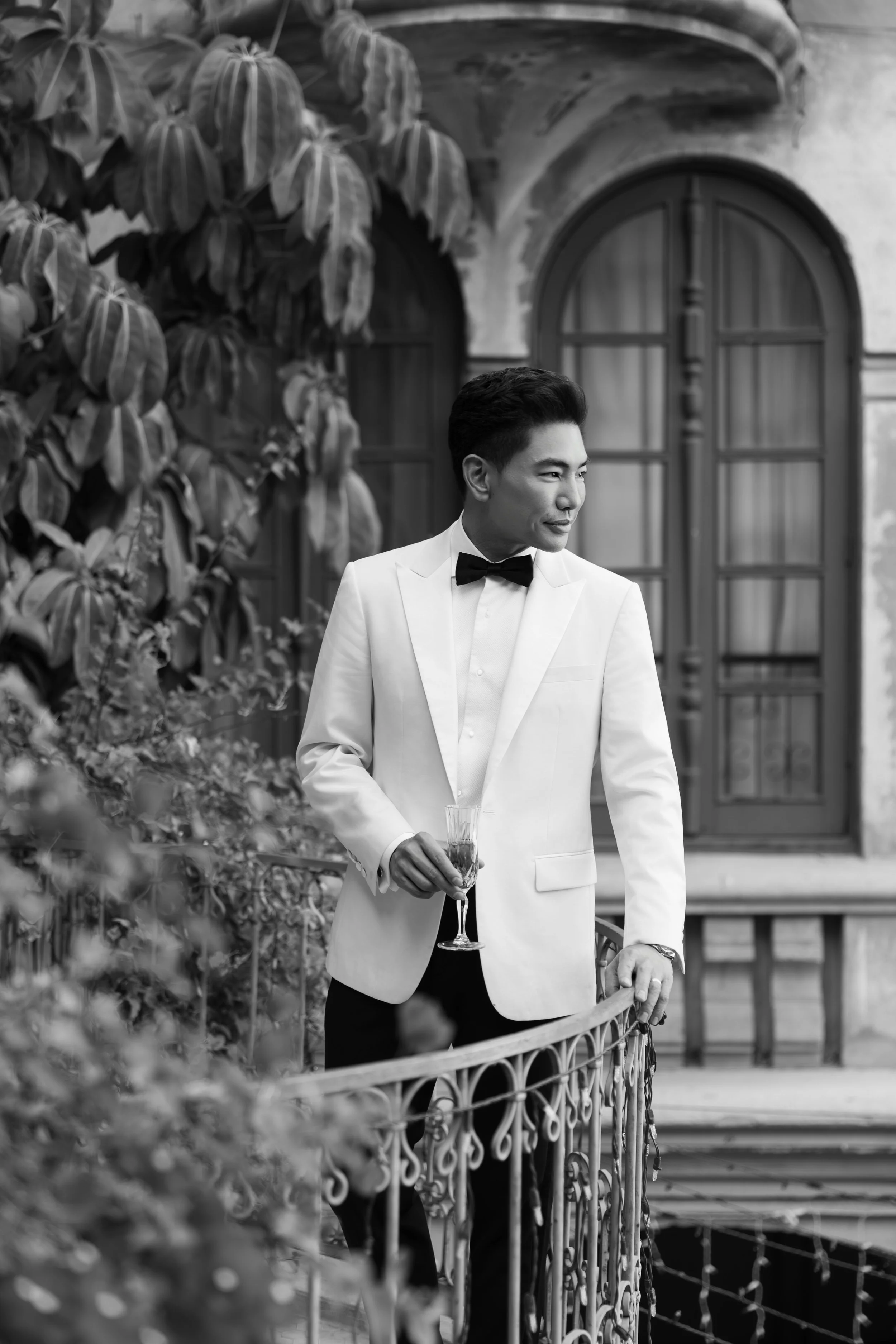 A man in a tuxedo with a bow tie holding a glass of wine, standing on a balcony with decorative railing, surrounded by plants and a building with arched windows.