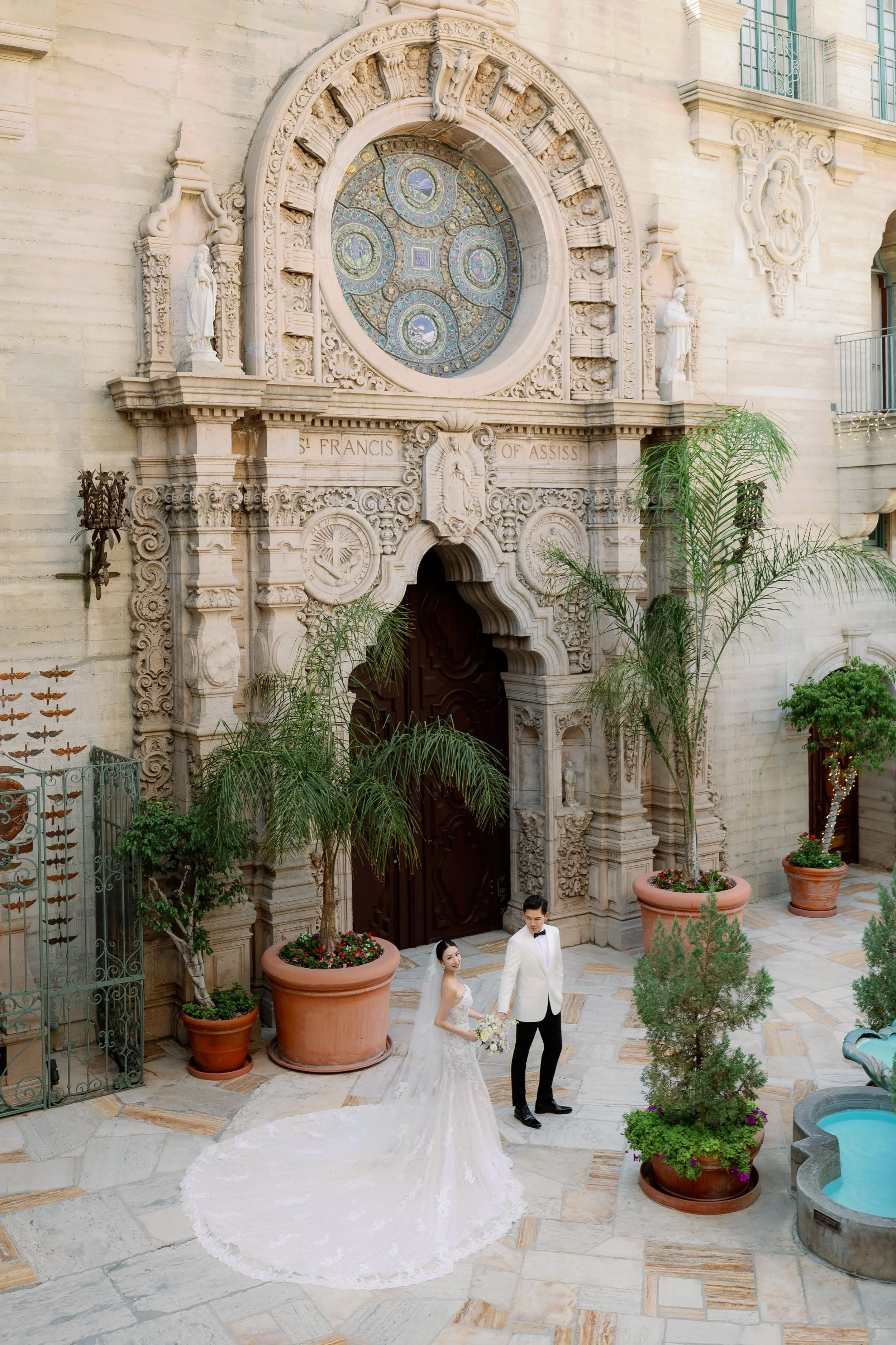 A bride and groom in wedding attire standing in a courtyard with ornate stone architecture, large potted plants, and a small fountain.