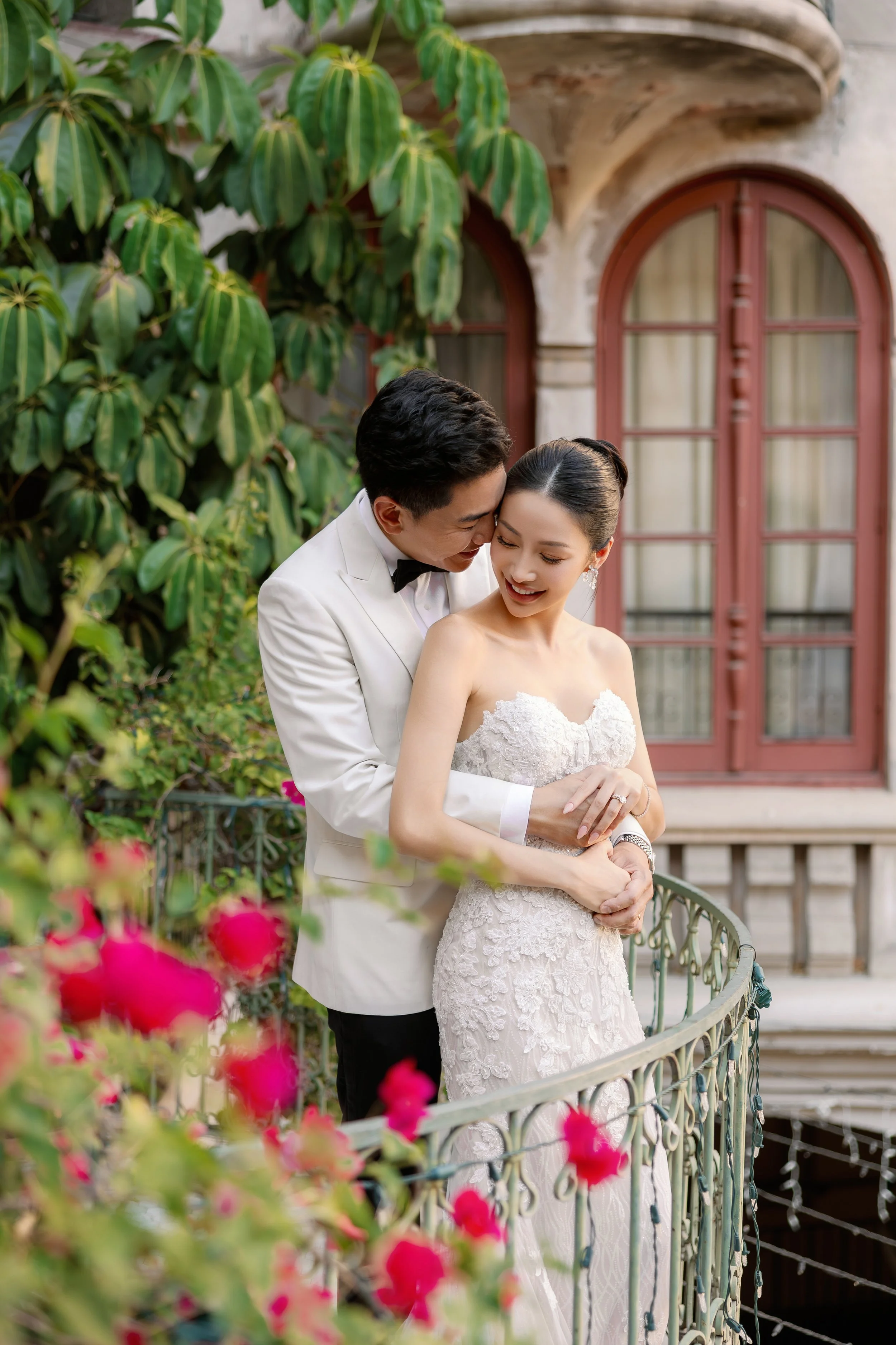 A bride and groom on a small balcony, sharing a joyful moment. The bride is in a white lace wedding dress, and the groom is in a white tuxedo with a black bow tie. There are pink flowers in the foreground and a green leafy background with a building 